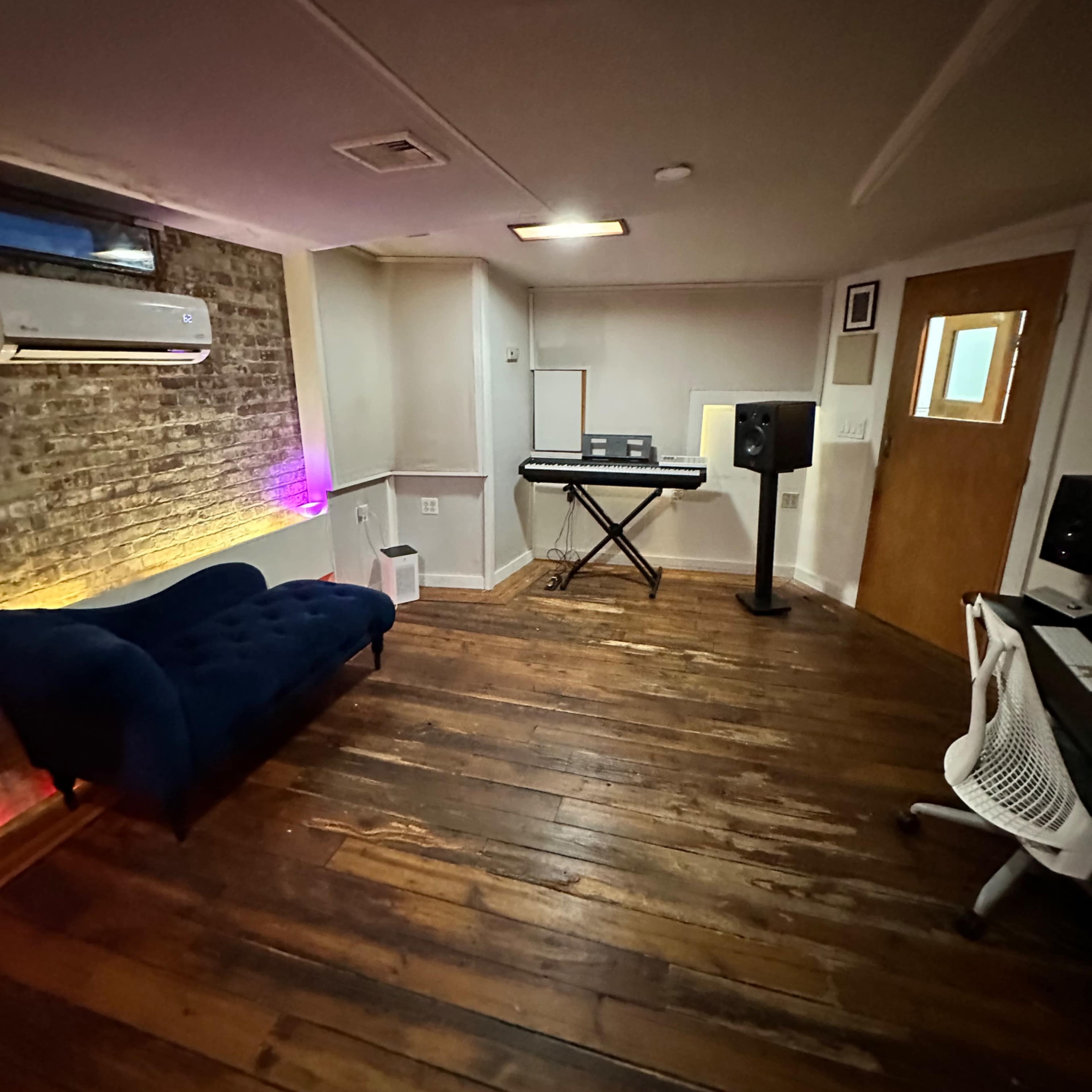 A room with wooden floors, a dark blue couch, a keyboard setup, a speaker, and a door leading to another space.
