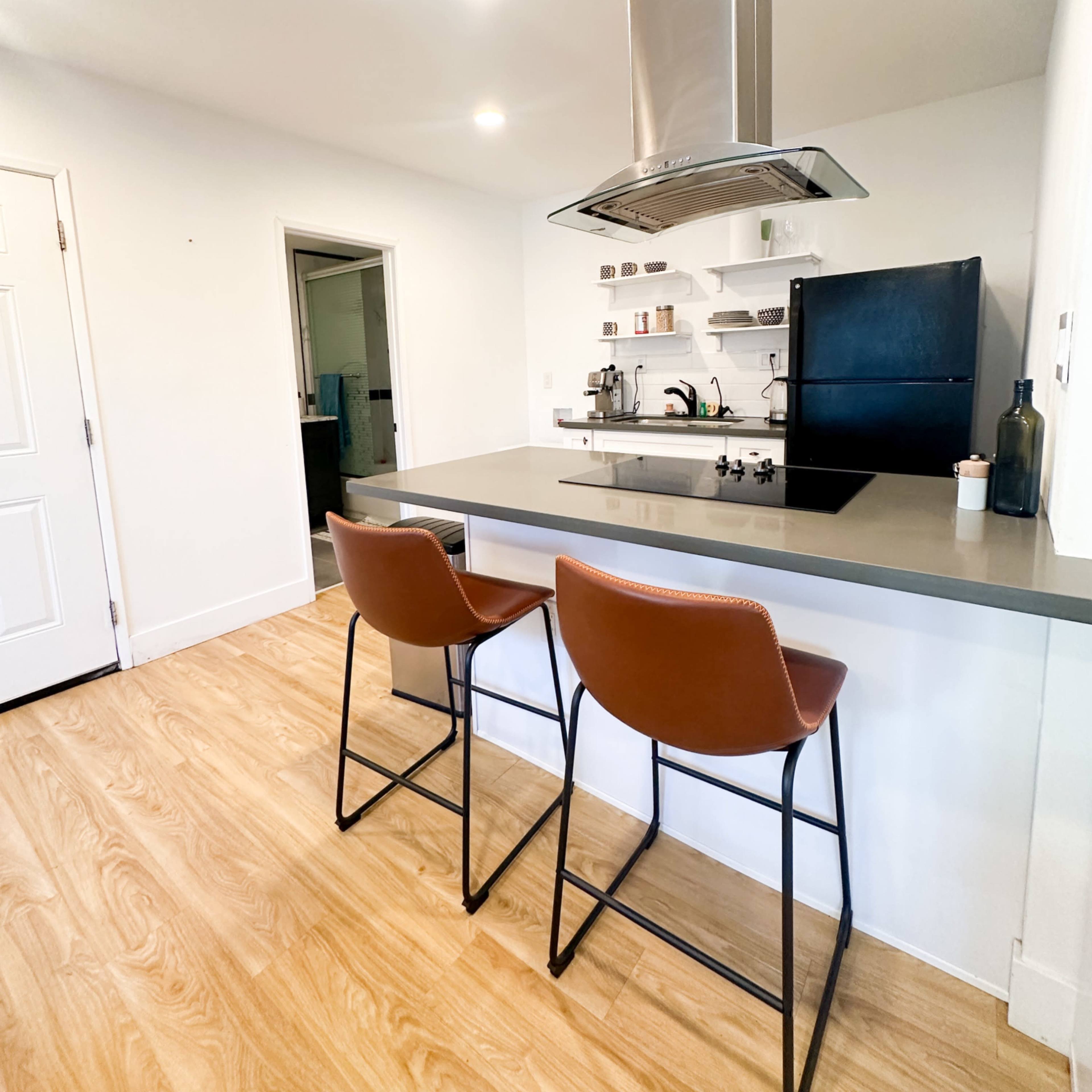 A modern kitchen features a central island with two leather bar stools, a black refrigerator, and a stainless steel range hood.