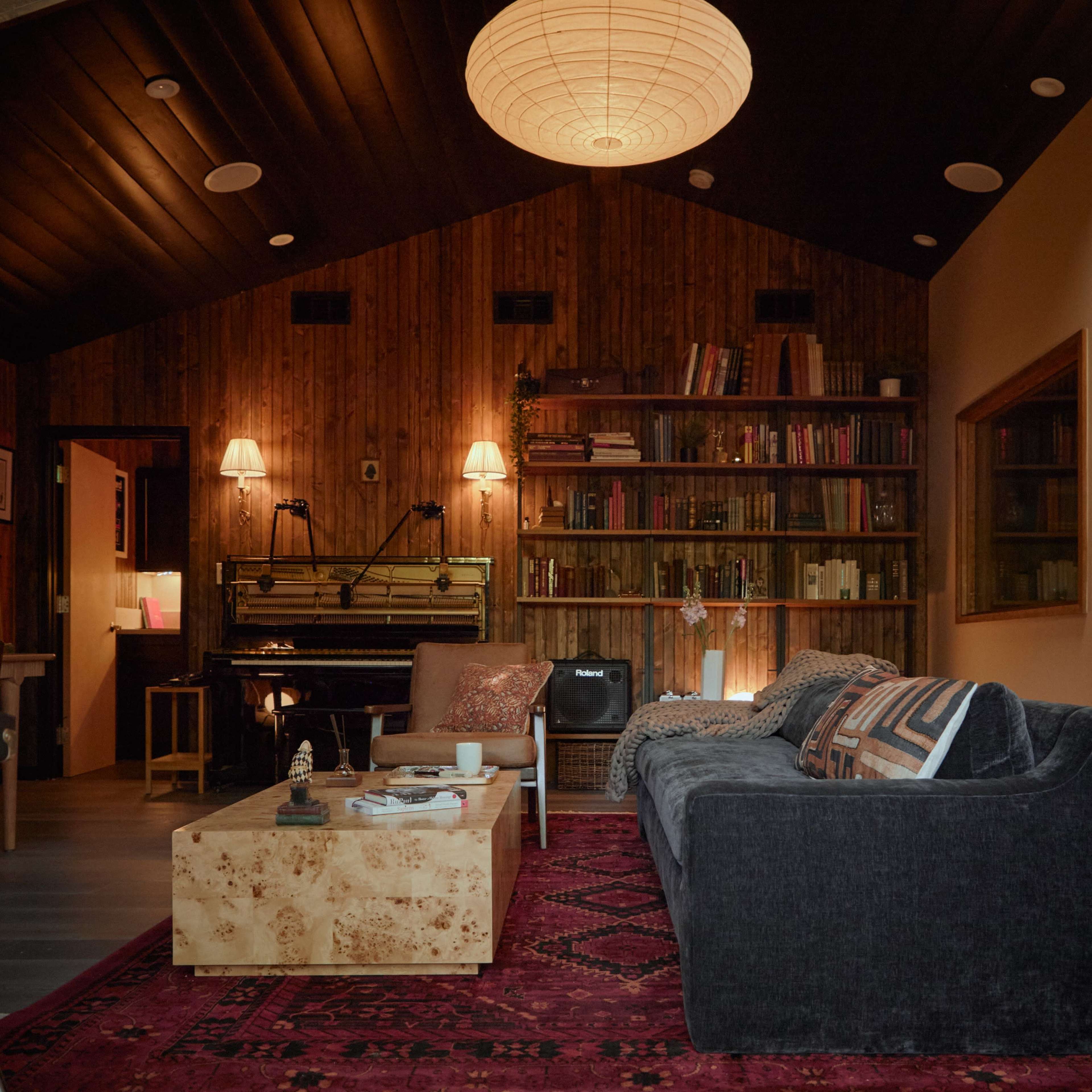 A cozy living room features a dark sectional sofa, a wooden bookshelf filled with books, a piano, and warm lighting from lamps and a hanging lantern.