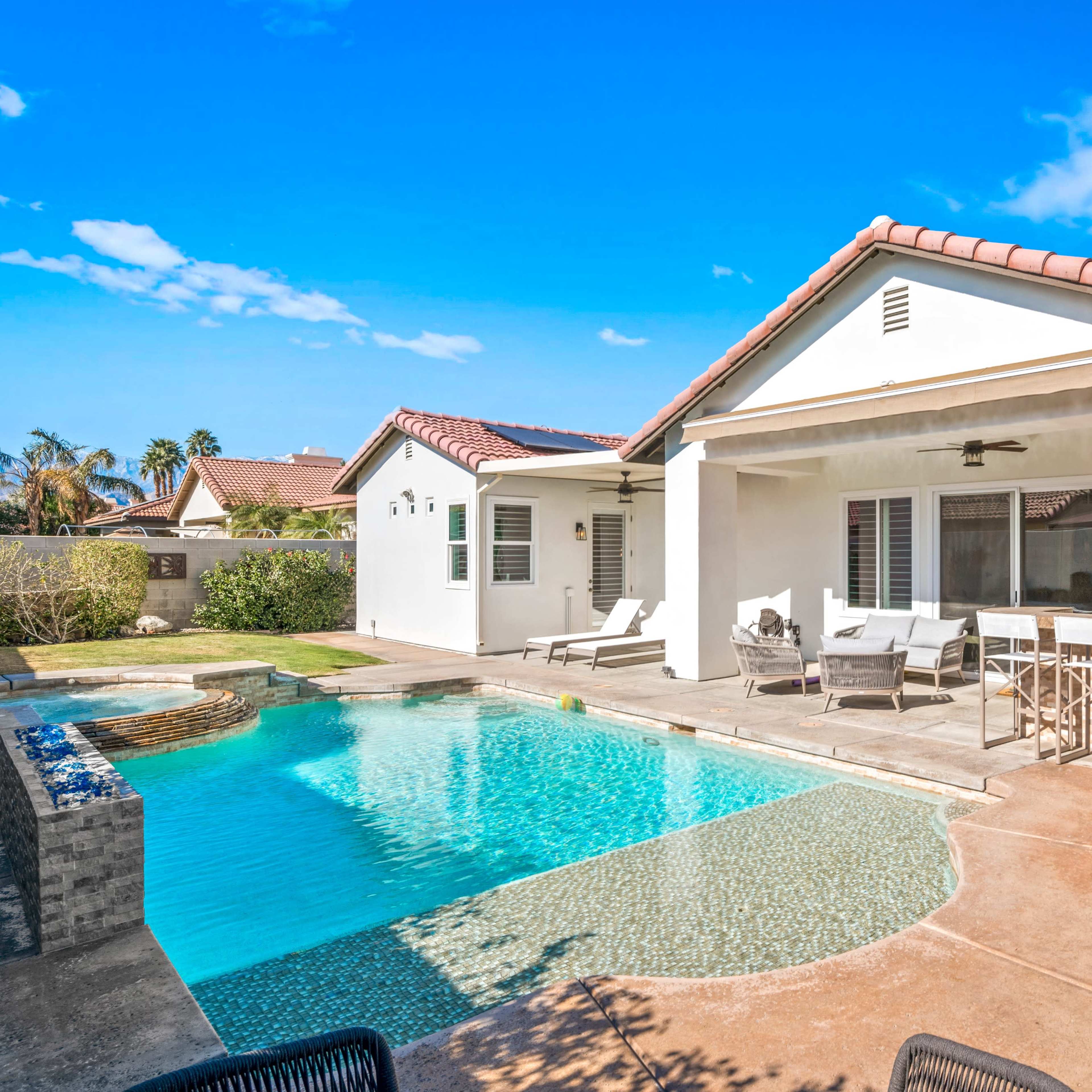 A modern home features a patio and a pool with a cascading water feature, surrounded by landscaped yard and clear blue skies.