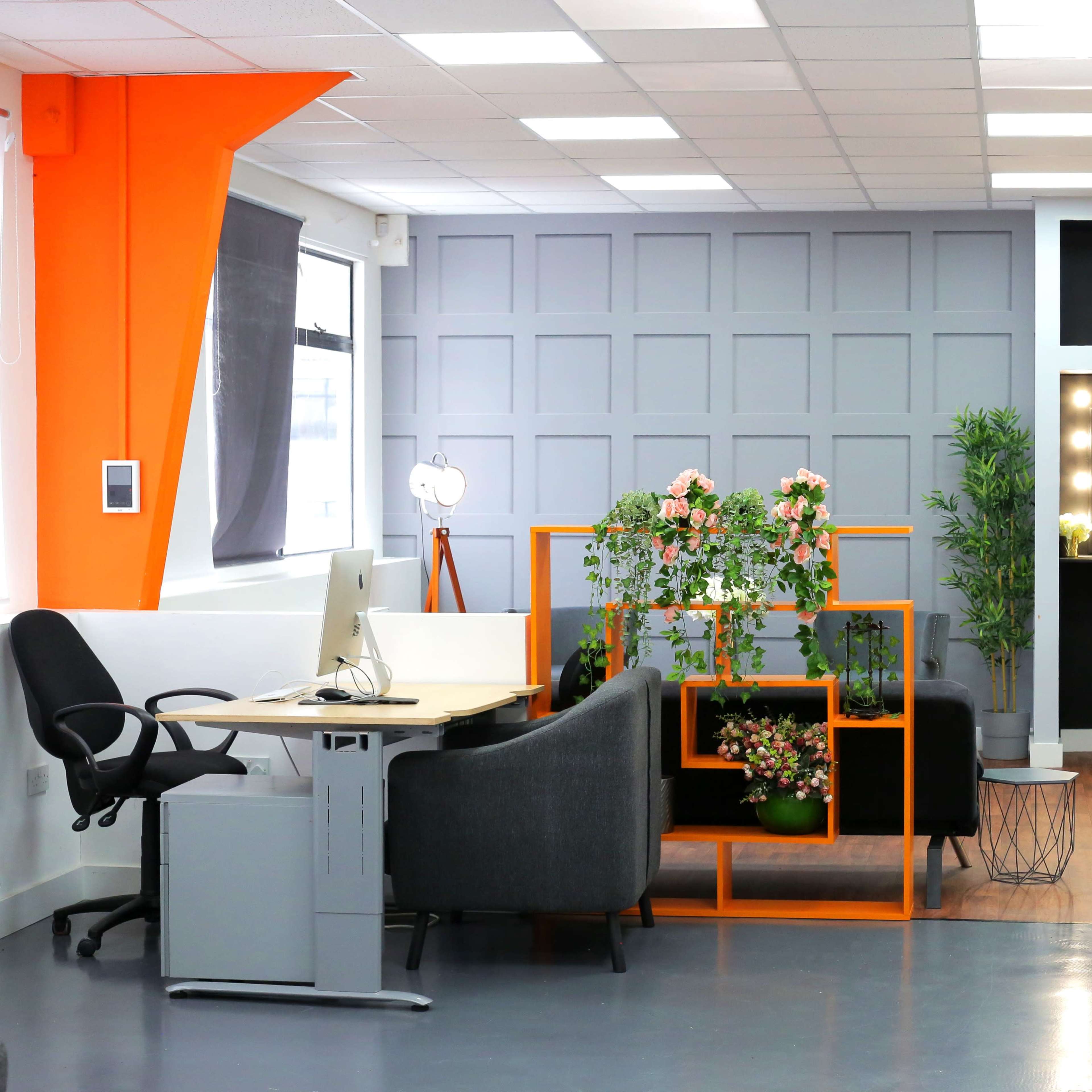 A modern office space featuring a desk with a computer, a seating area, decorative plants, and a vanity mirror area in the background.