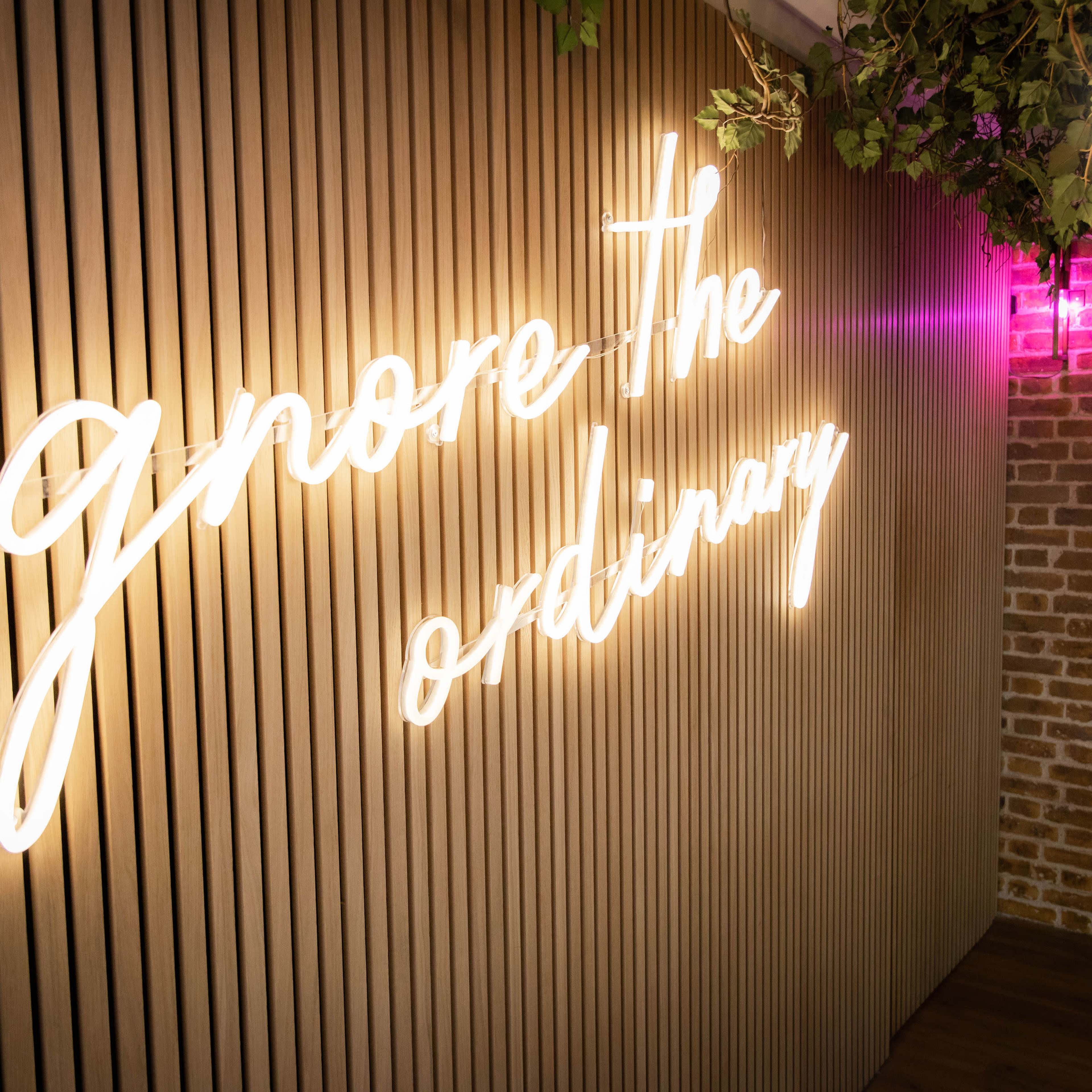 A neon sign that reads "ignore the ordinary" is illuminated on a wooden slat wall with greenery overhead.