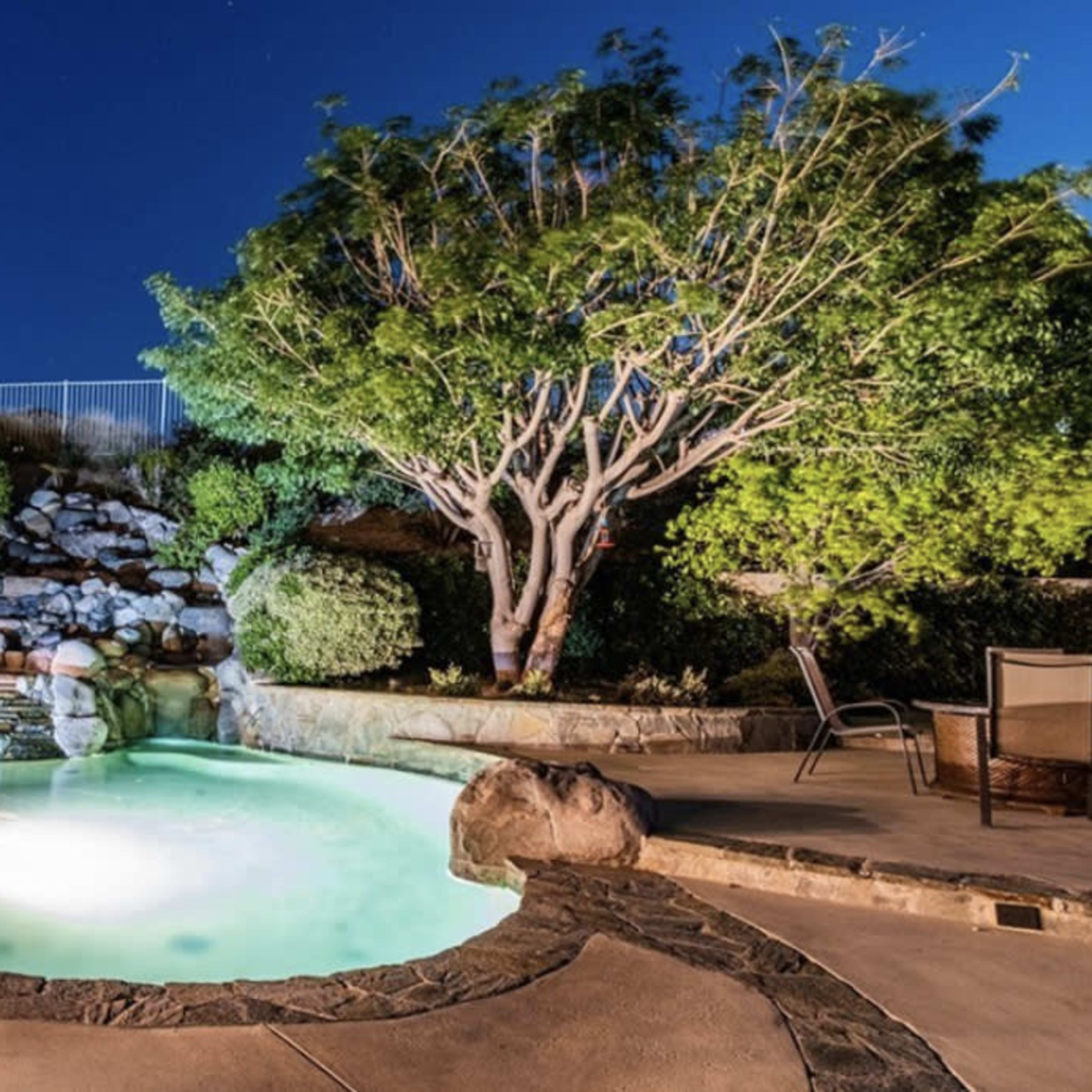 A backyard pool with a waterfall and landscaping is illuminated at night under a clear sky.