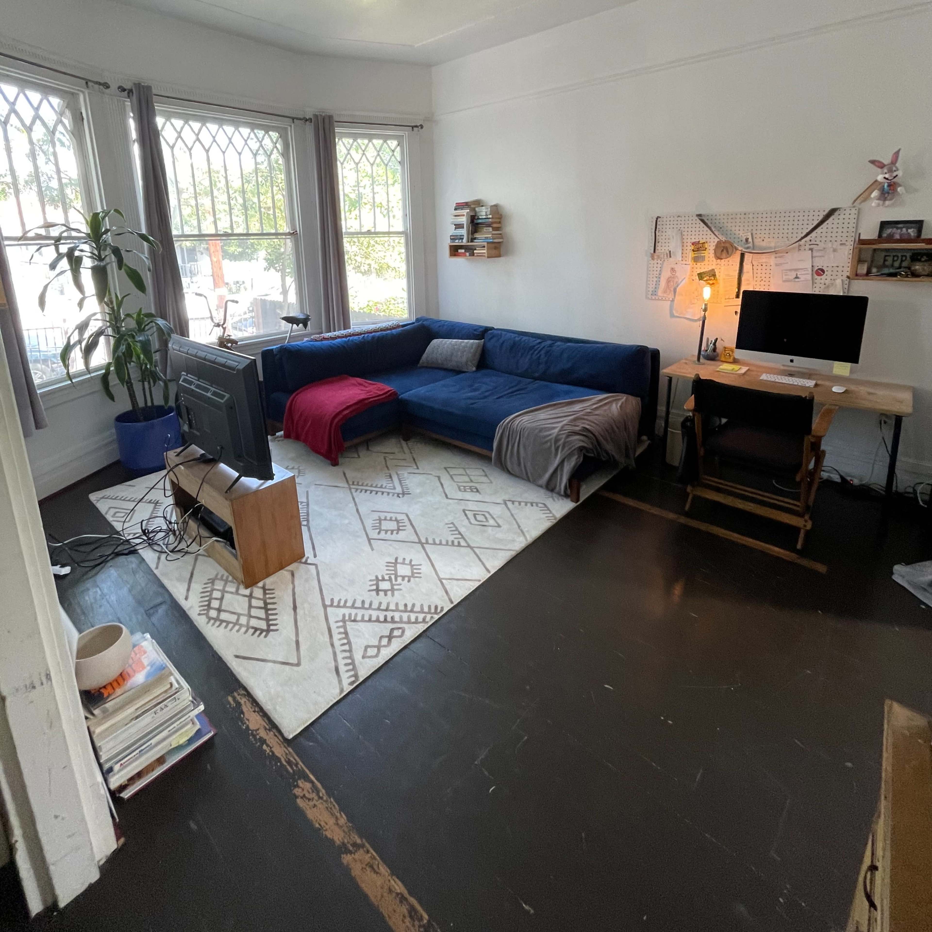 A cozy living room with a blue sectional sofa, a small desk with a computer, and a large window allowing natural light to enter the space.