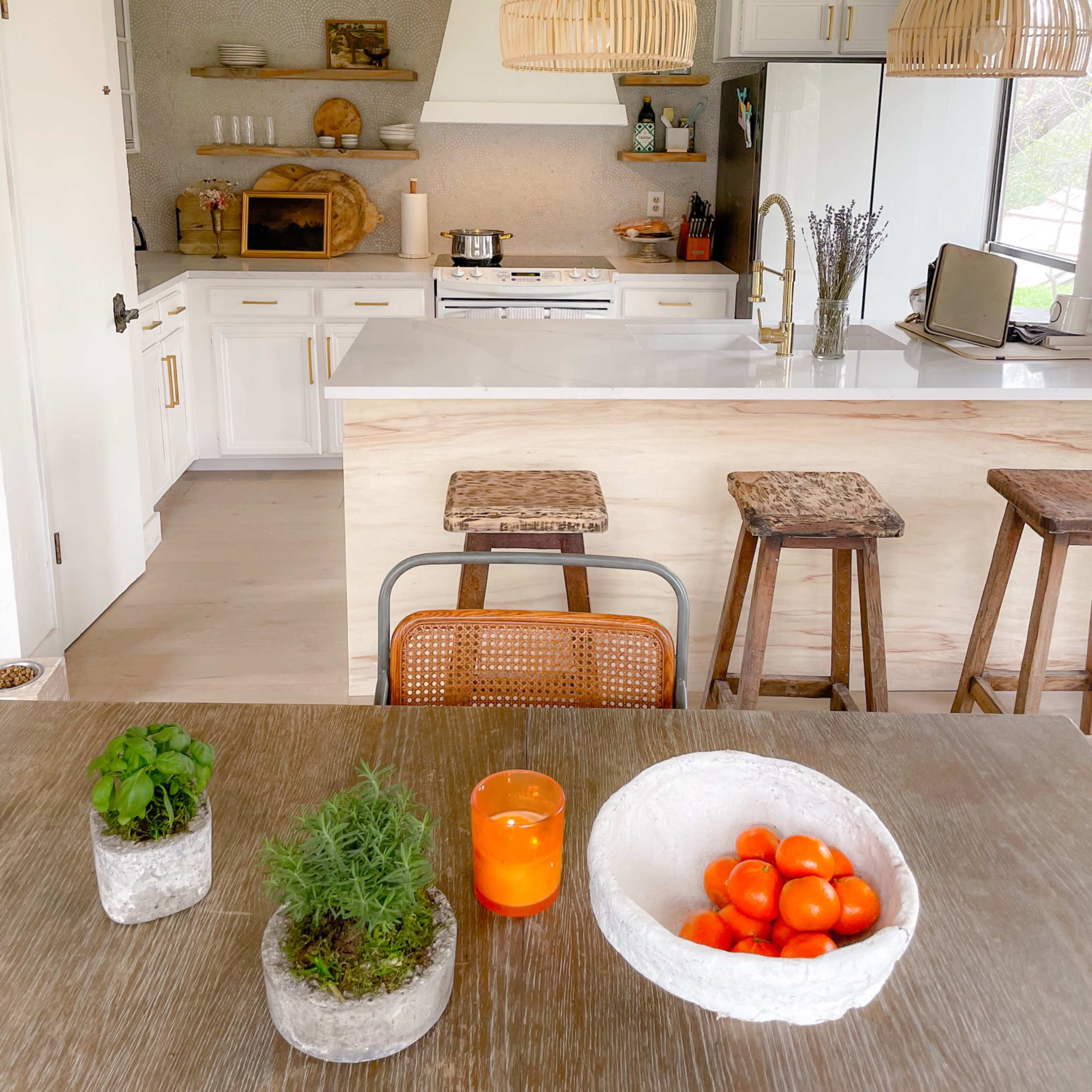 A modern kitchen featuring white cabinetry, a central island with bar stools, and decorative elements like potted herbs and a bowl of oranges on the dining table.