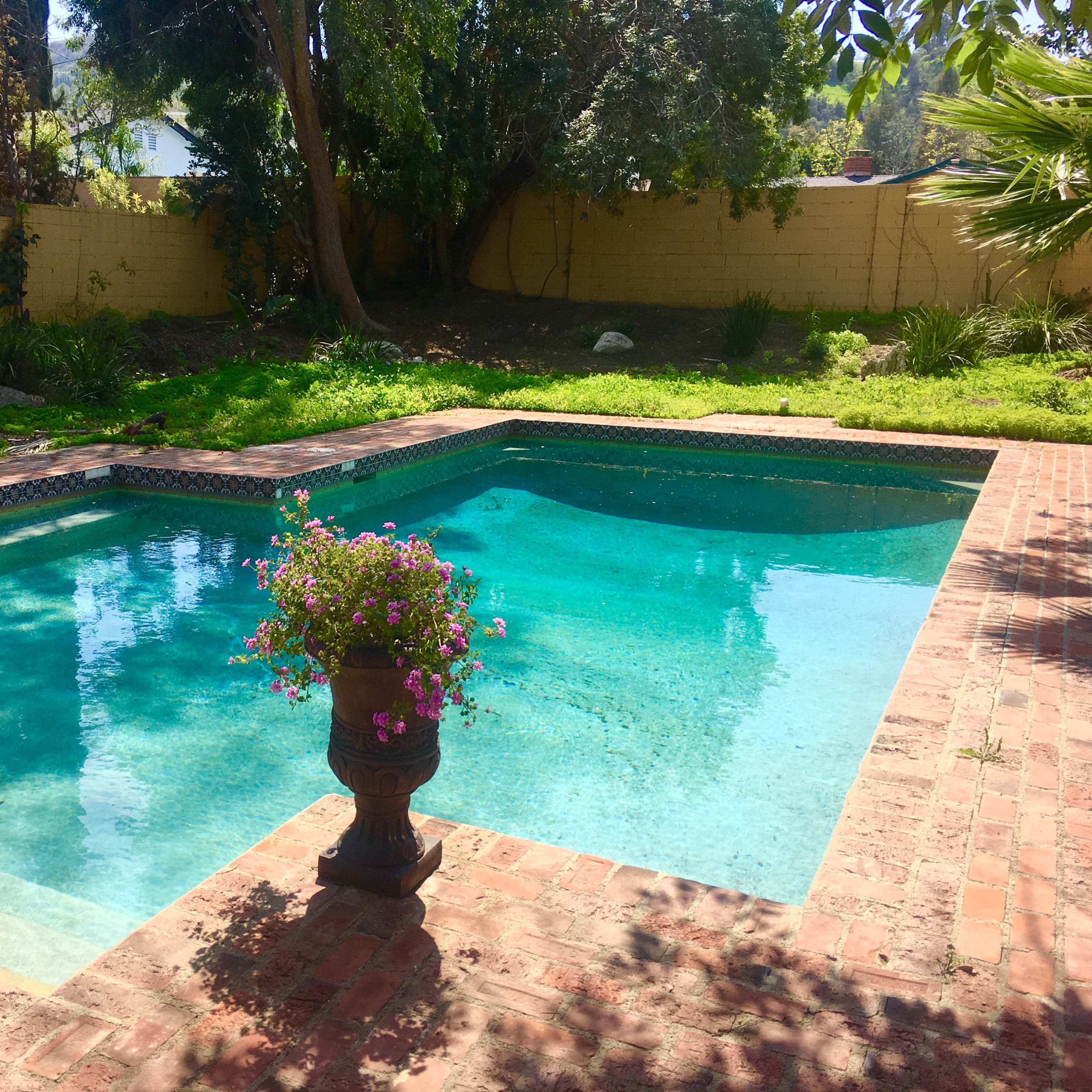 A swimming pool with clear blue water is surrounded by a brick patio and a decorative planter with flowers in a garden setting.