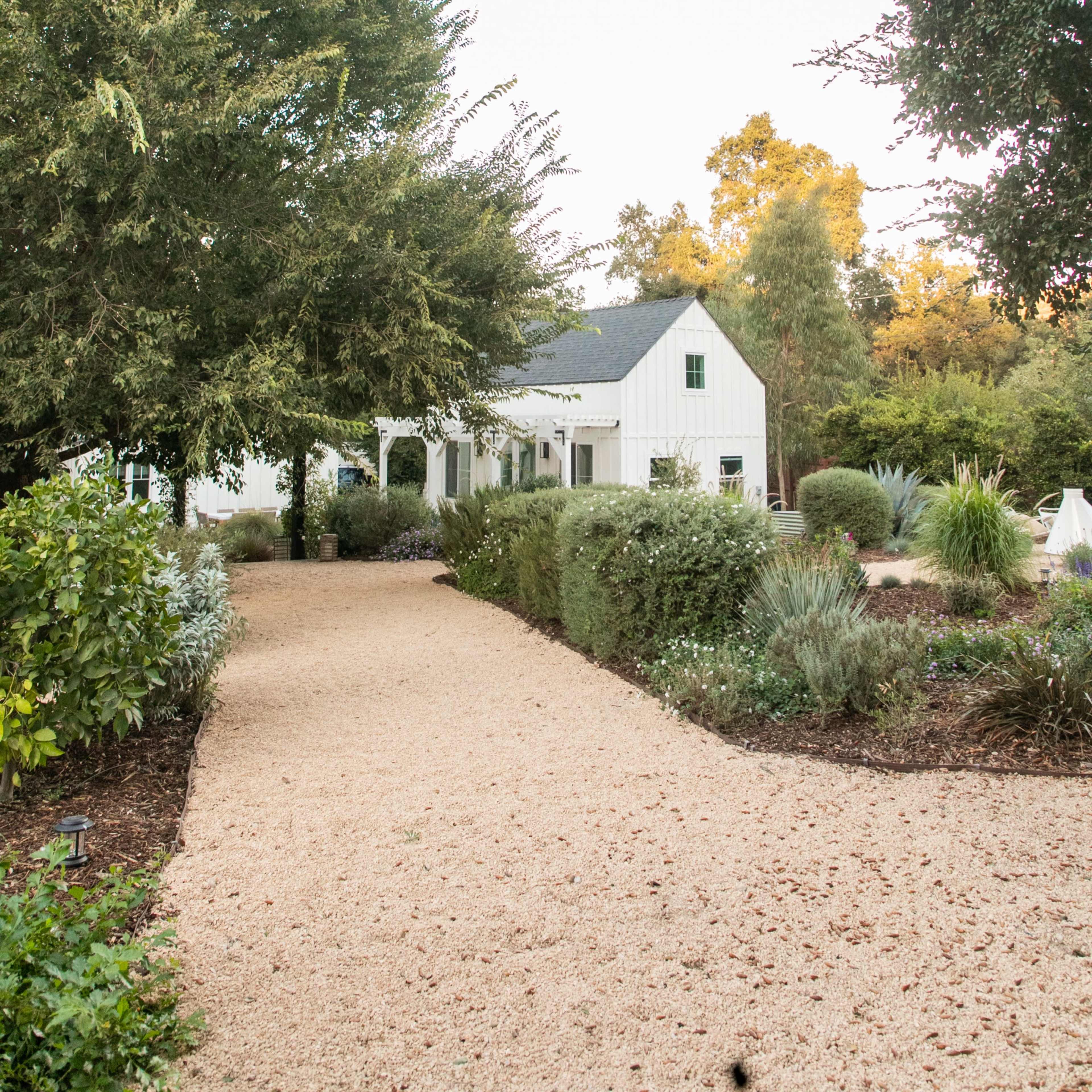 A gravel pathway leads to a white farmhouse surrounded by trees and well-maintained landscaping.