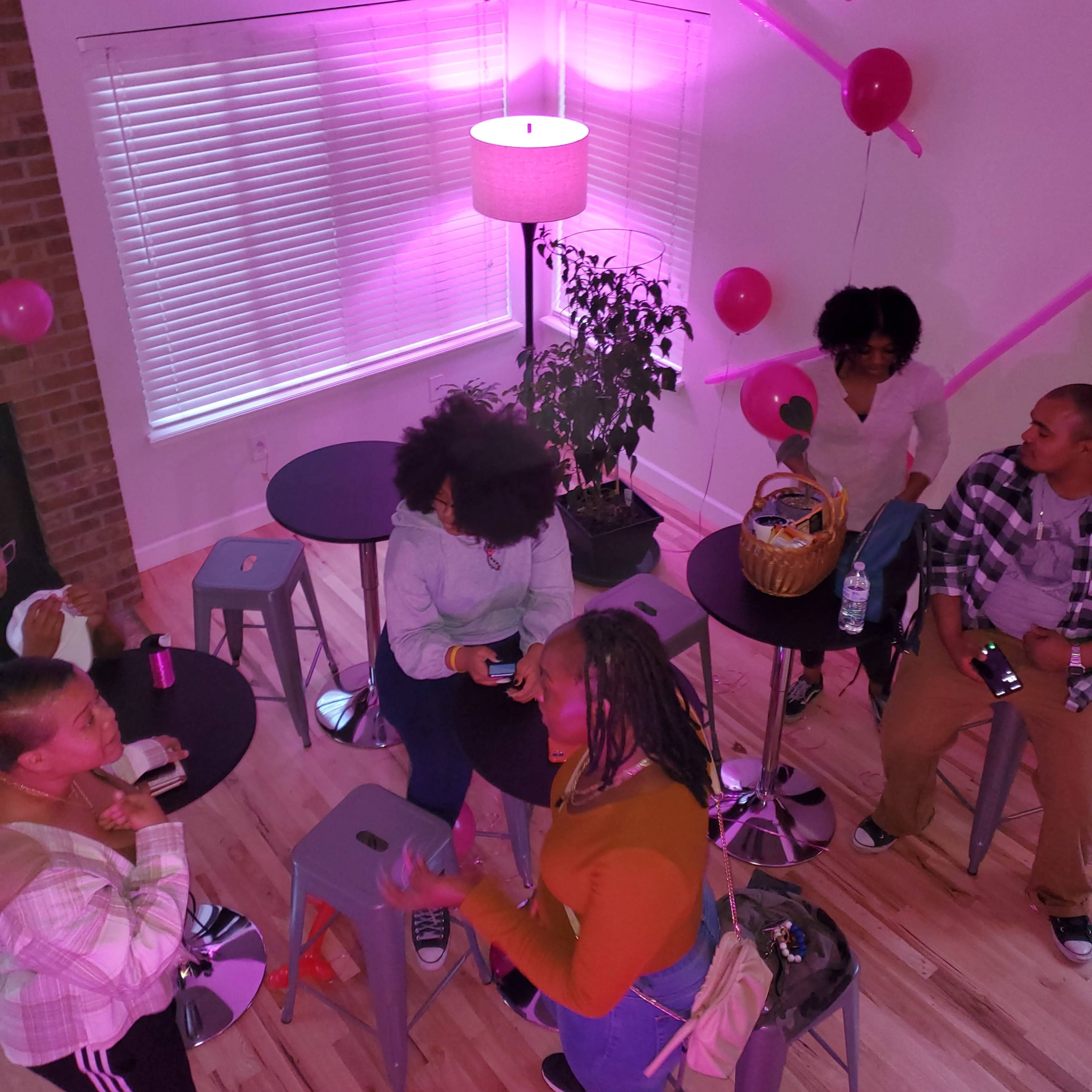 A group of people chats around small tables in a brightly lit room decorated with pink balloons and a plant.