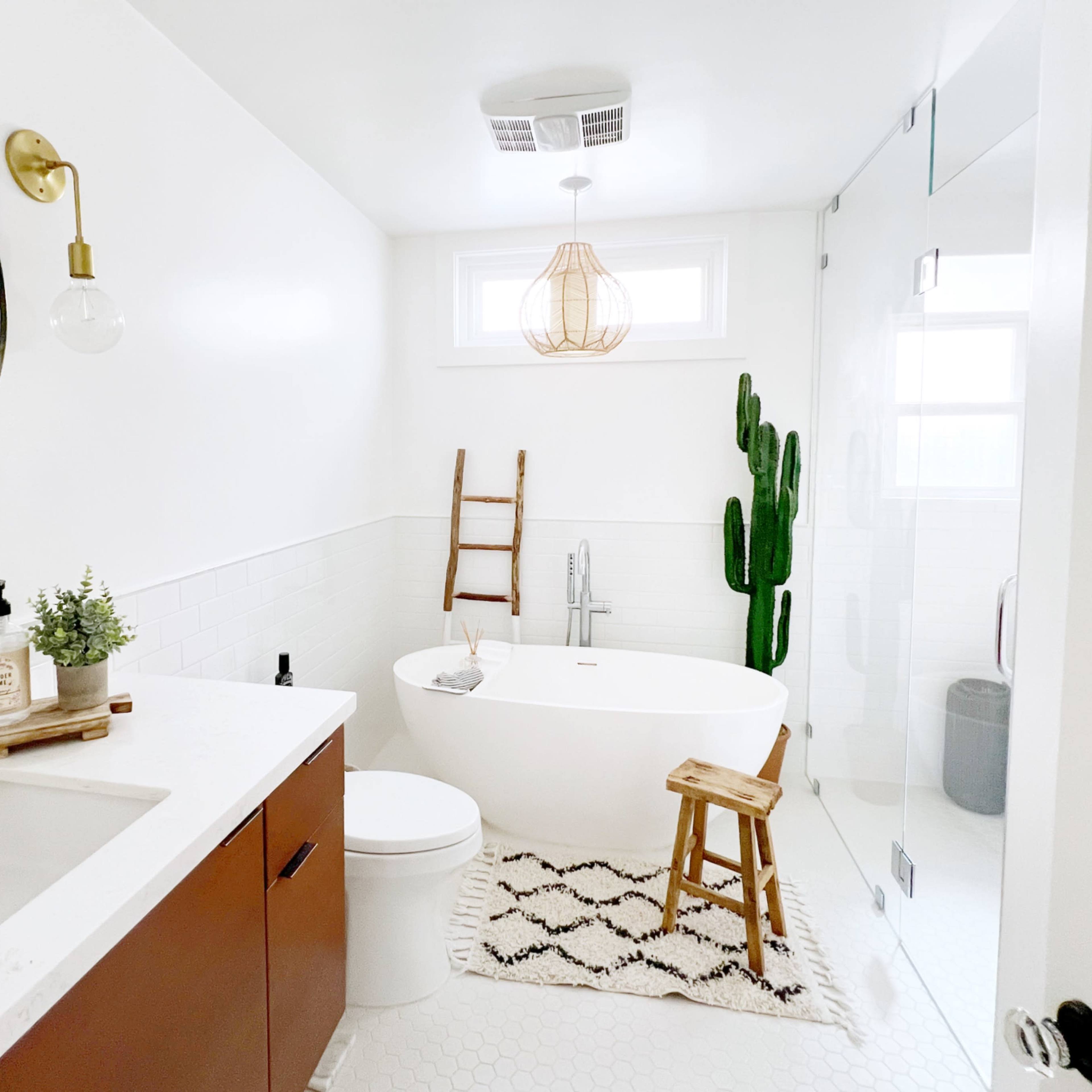 The bathroom features a freestanding bathtub, a modern sink with wooden cabinetry, and a tall cactus next to a ladder, all set against a clean white backdrop.