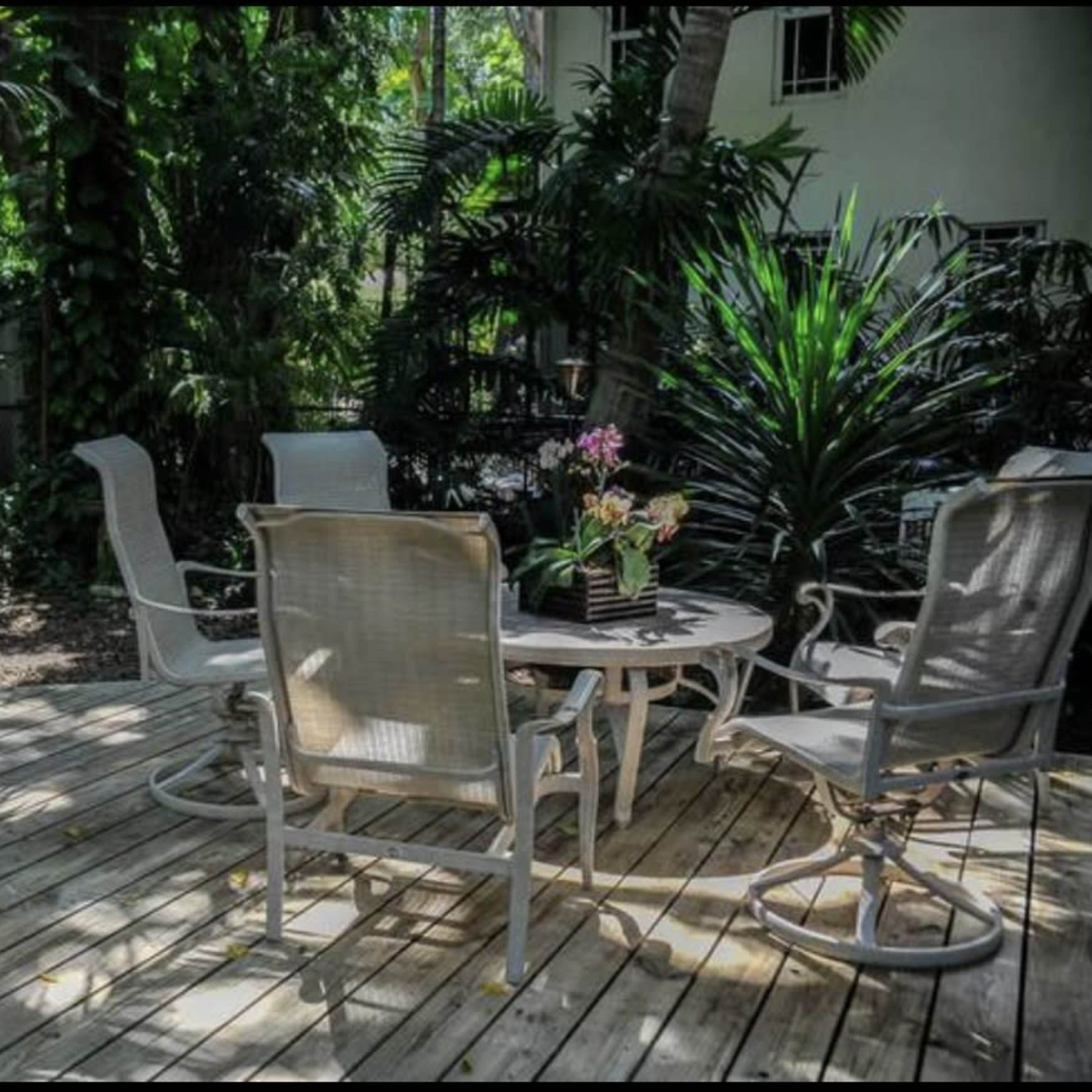 A wooden deck with four white chairs and a round table is surrounded by lush greenery and potted plants.