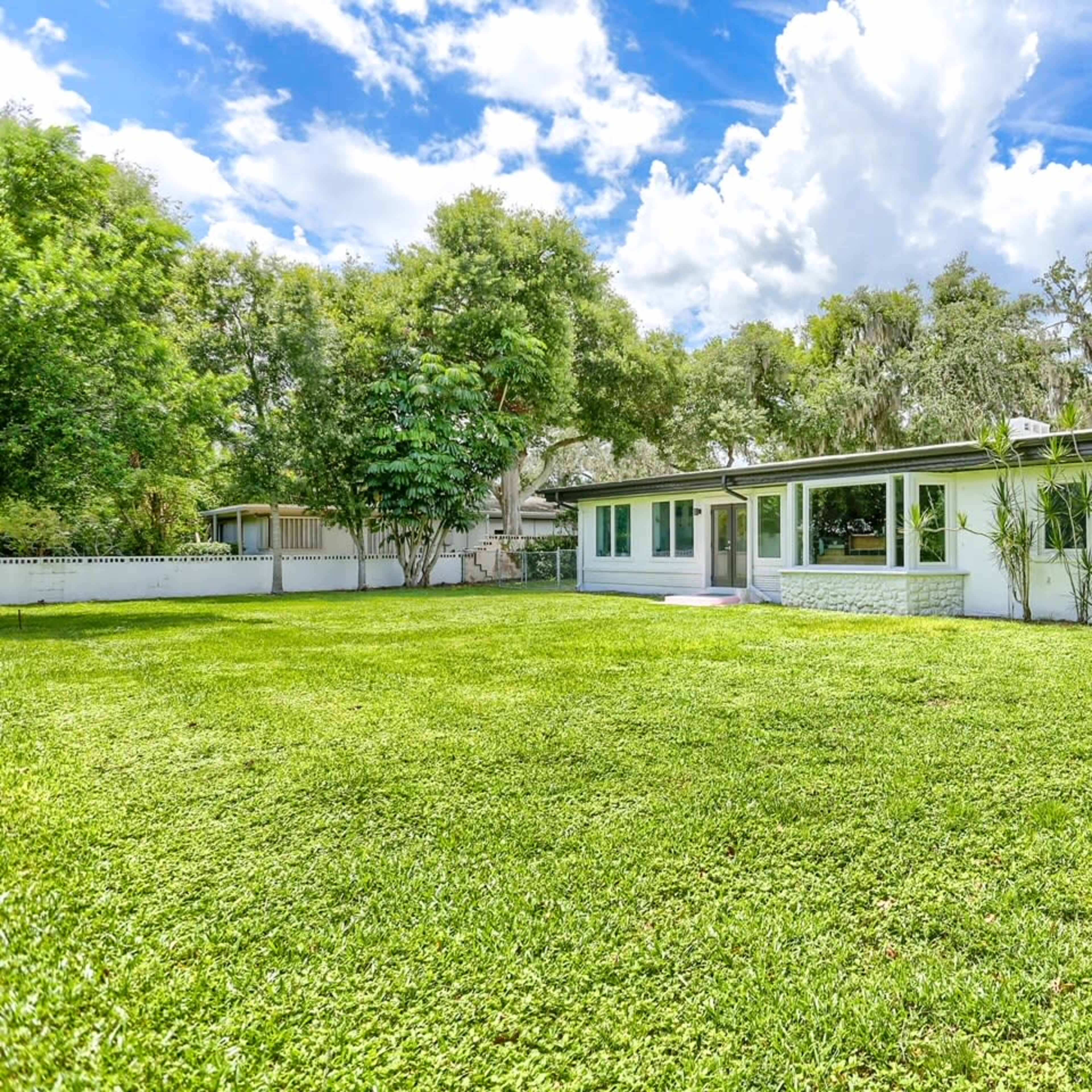 A single-story house with a large green lawn, surrounded by trees and under a blue sky with clouds.