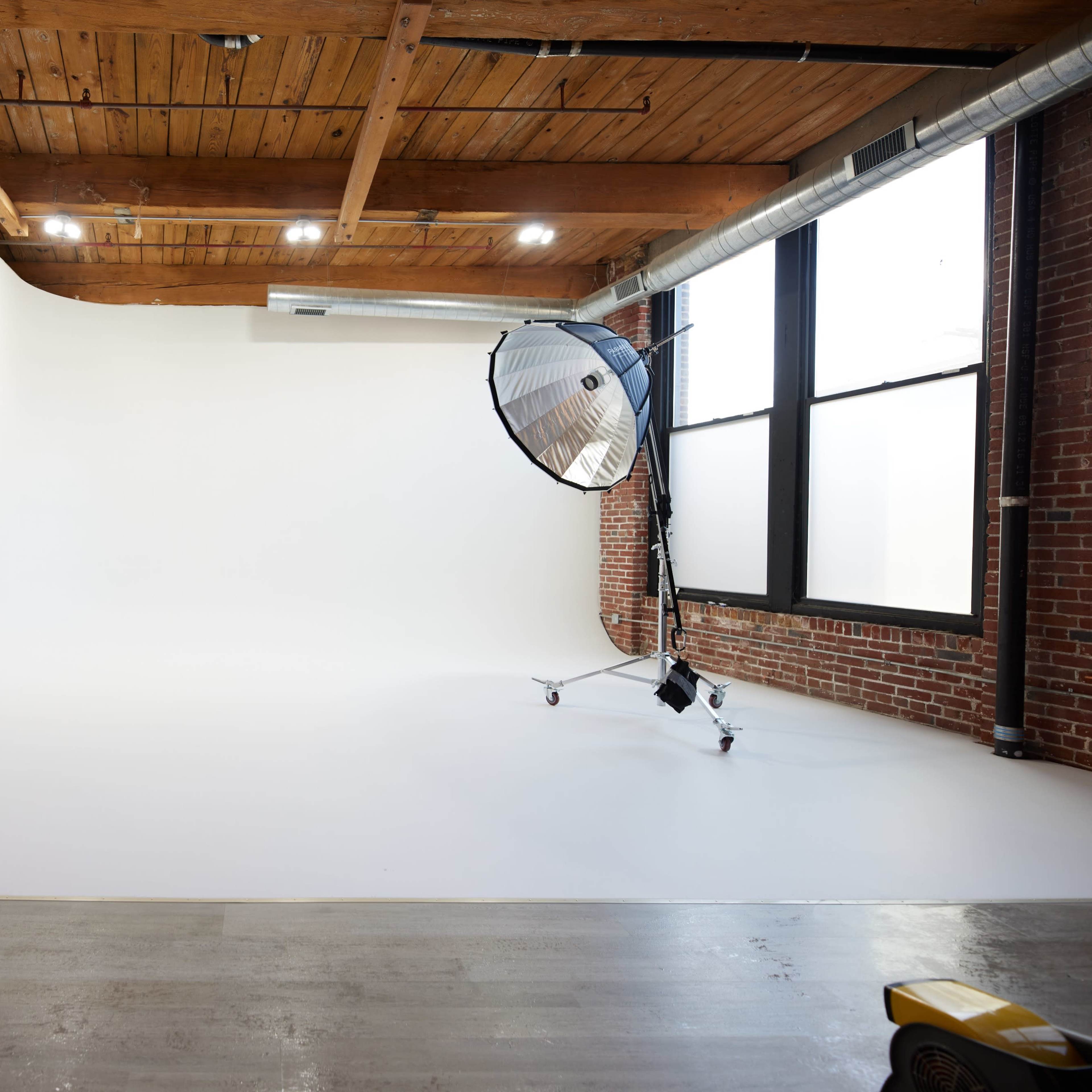 A photography studio with a white backdrop, a large softbox light on wheels, and exposed wooden beams overhead.