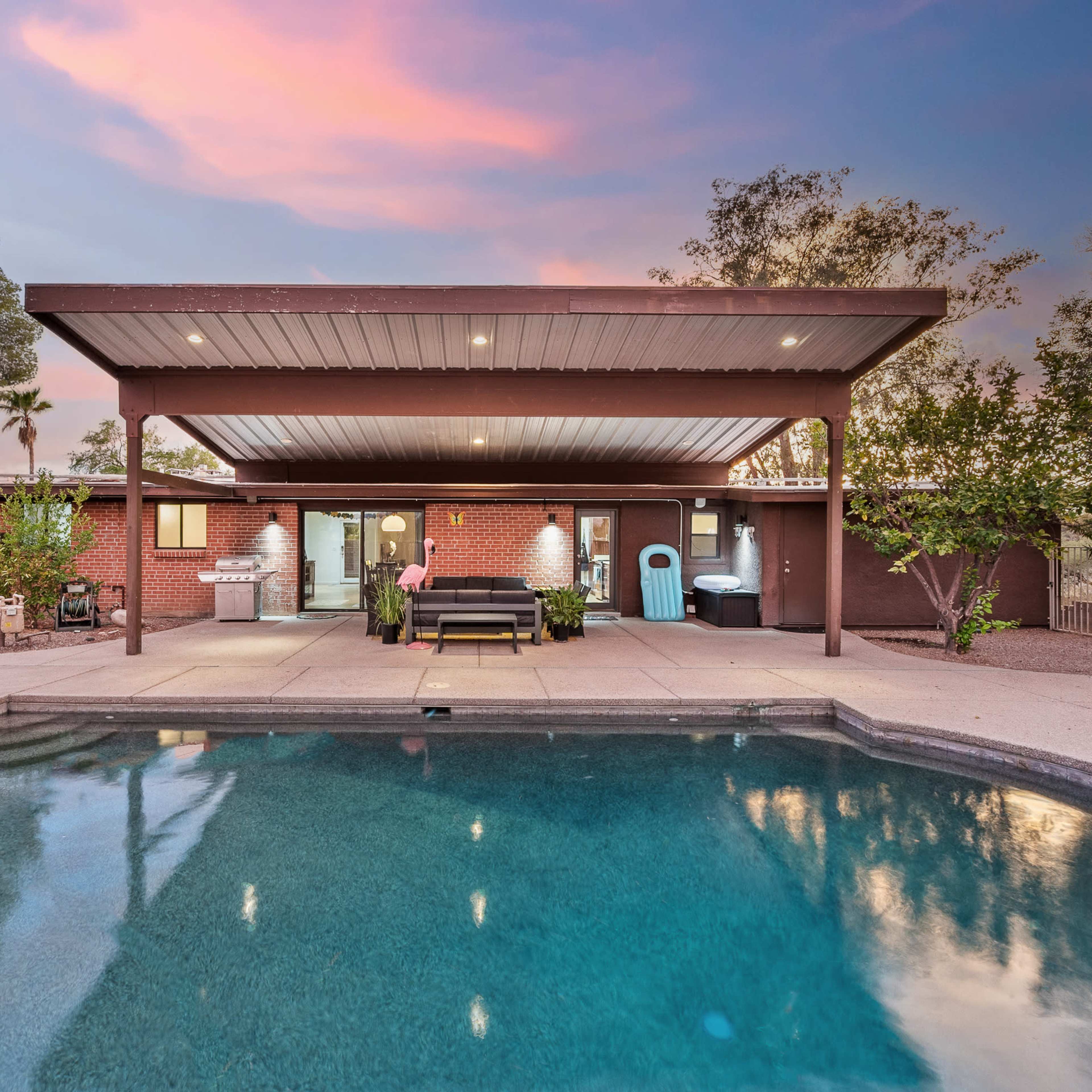 A patio area with a swimming pool is shown behind a house, featuring a covered seating space and landscaped plants under a colorful sunset sky.
