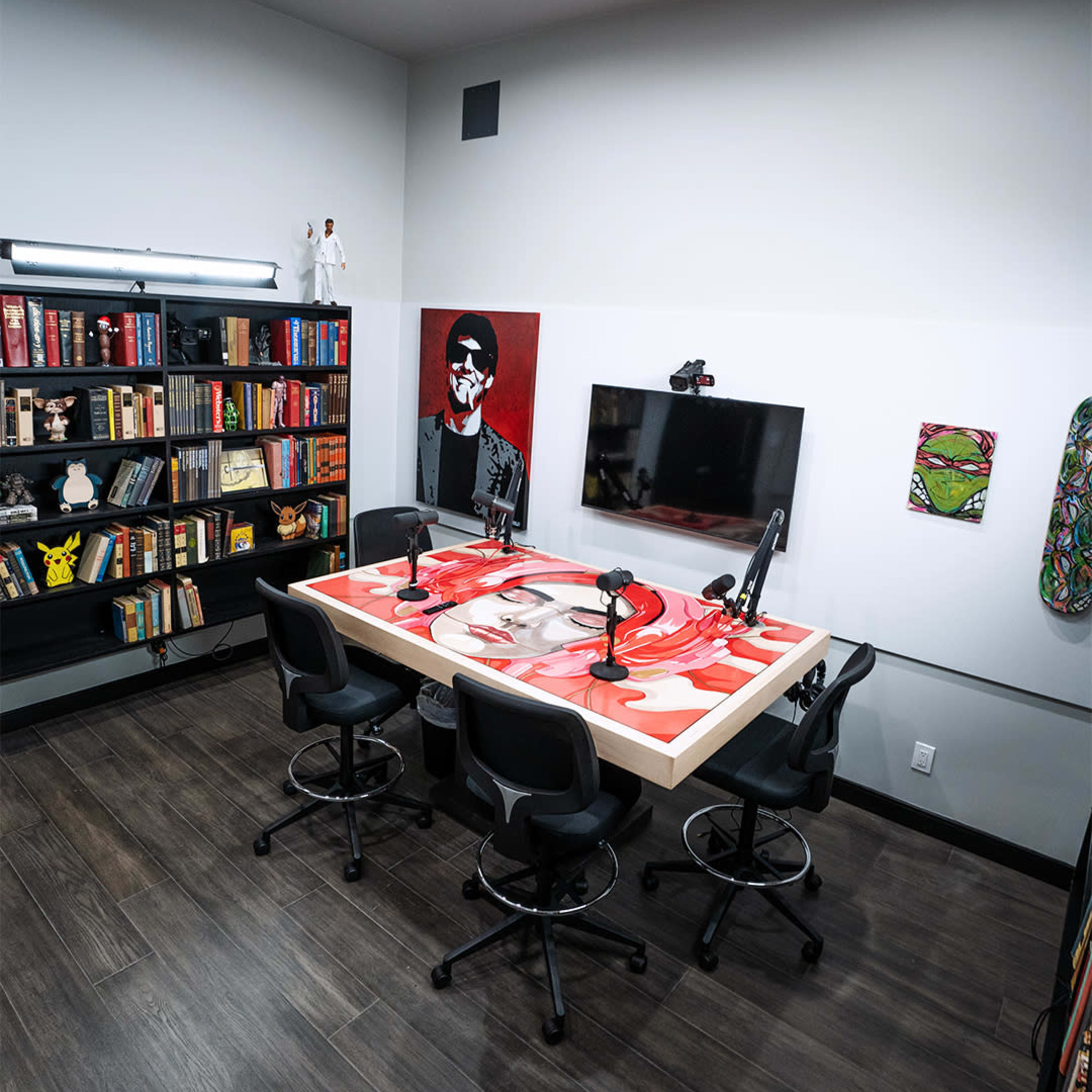 A modern podcast studio with a large table, four microphone setups, a wall-mounted television, and bookshelves filled with various books and decor.