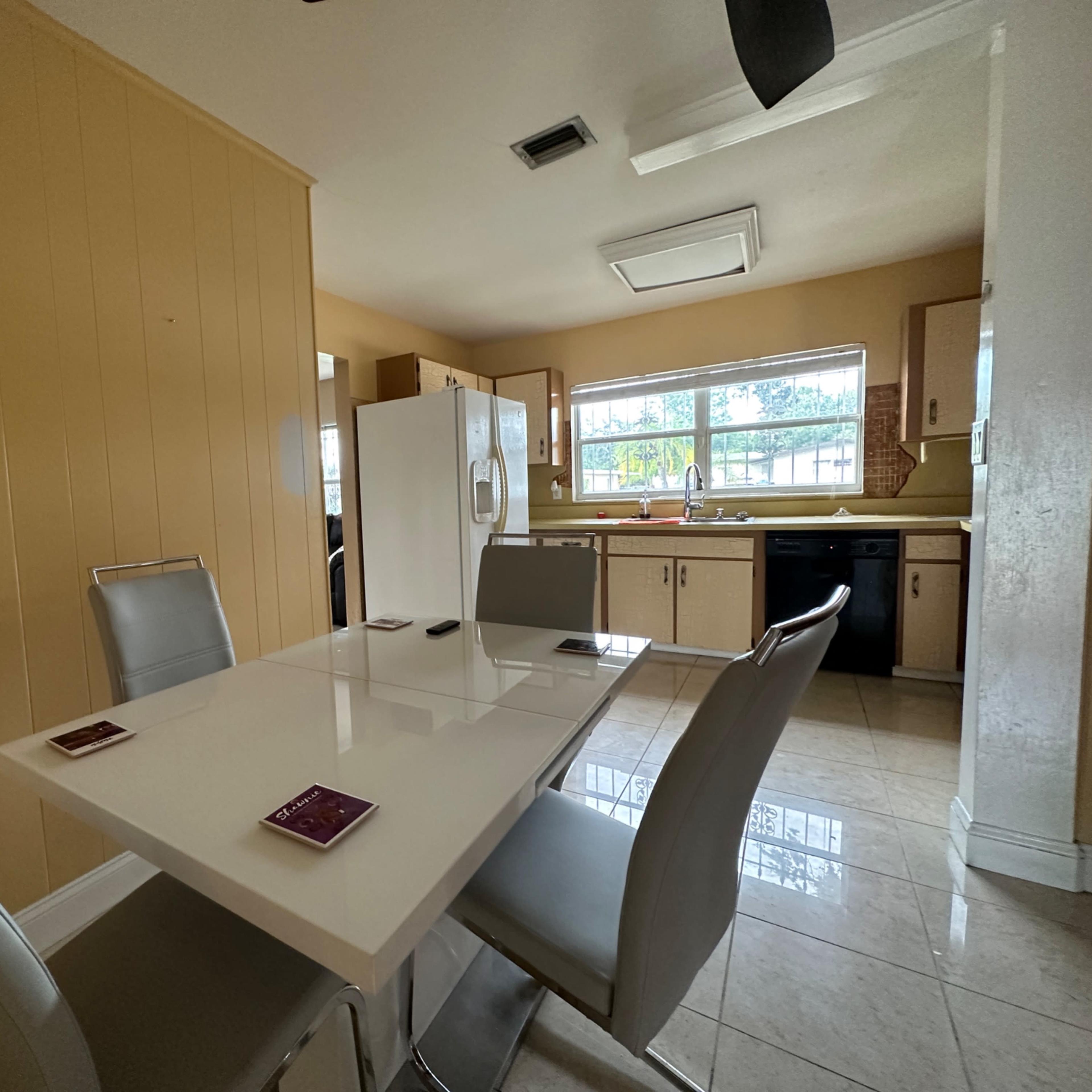 A modern kitchen features a light-colored table surrounded by four chairs, with appliances and cabinets visible in the background.