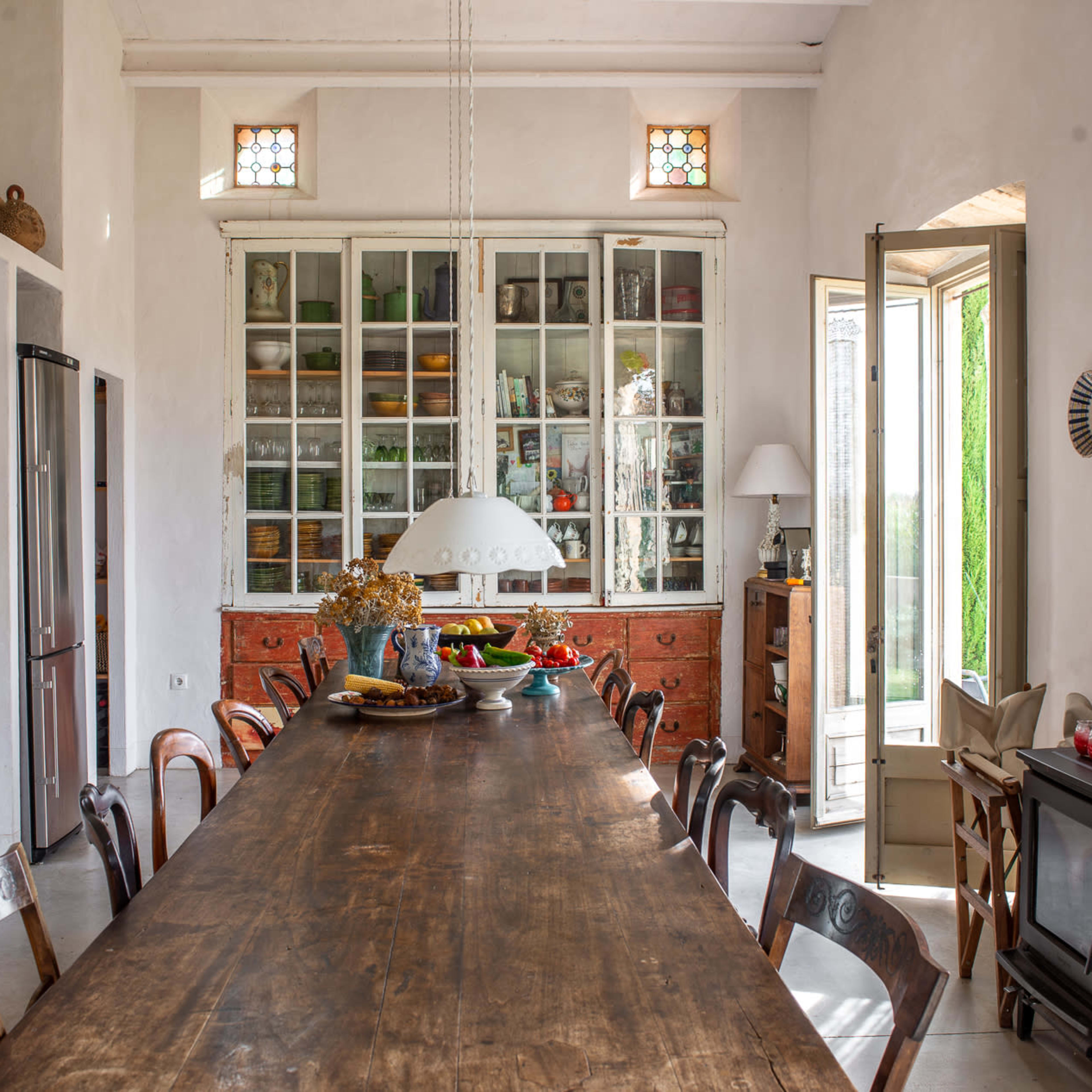 A spacious kitchen with a long wooden table, large glass cabinets displaying various dishes, and an open door leading to an outdoor area.