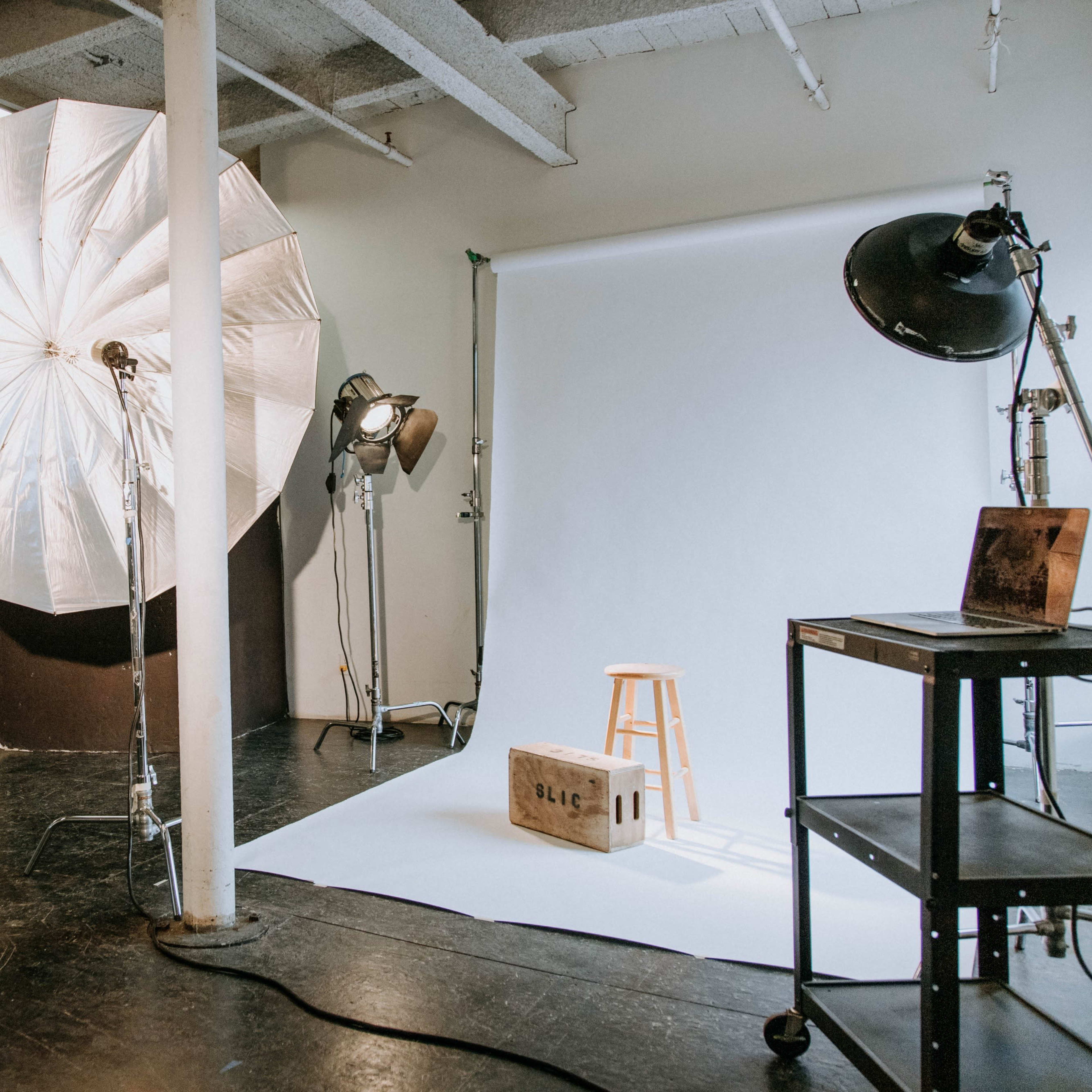 A photography studio with a large umbrella light, a backdrop, a stool, and several lighting setups.