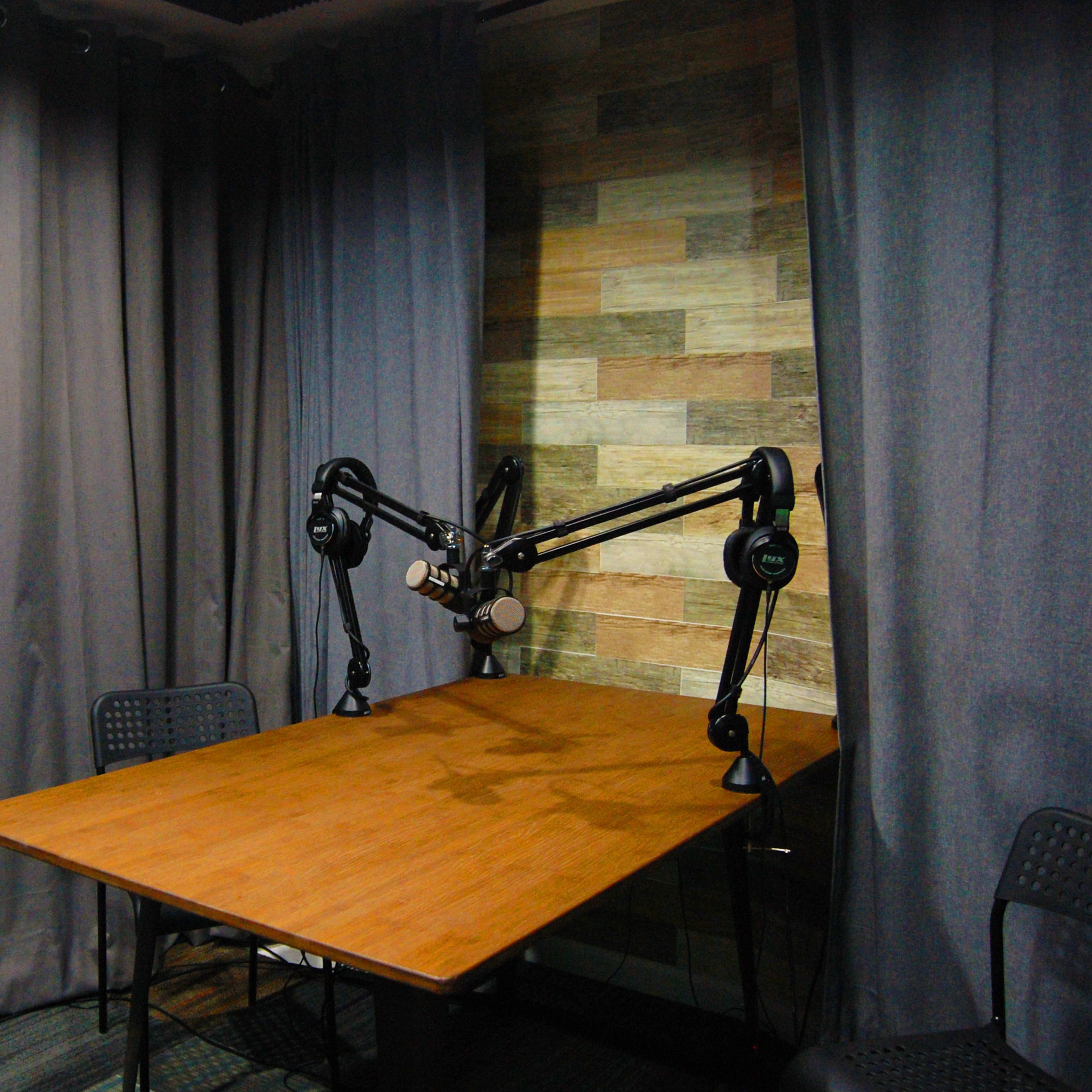 A small recording studio features a wooden table, two black chairs, and microphones mounted on adjustable arms, surrounded by dark curtains and a patterned wall.