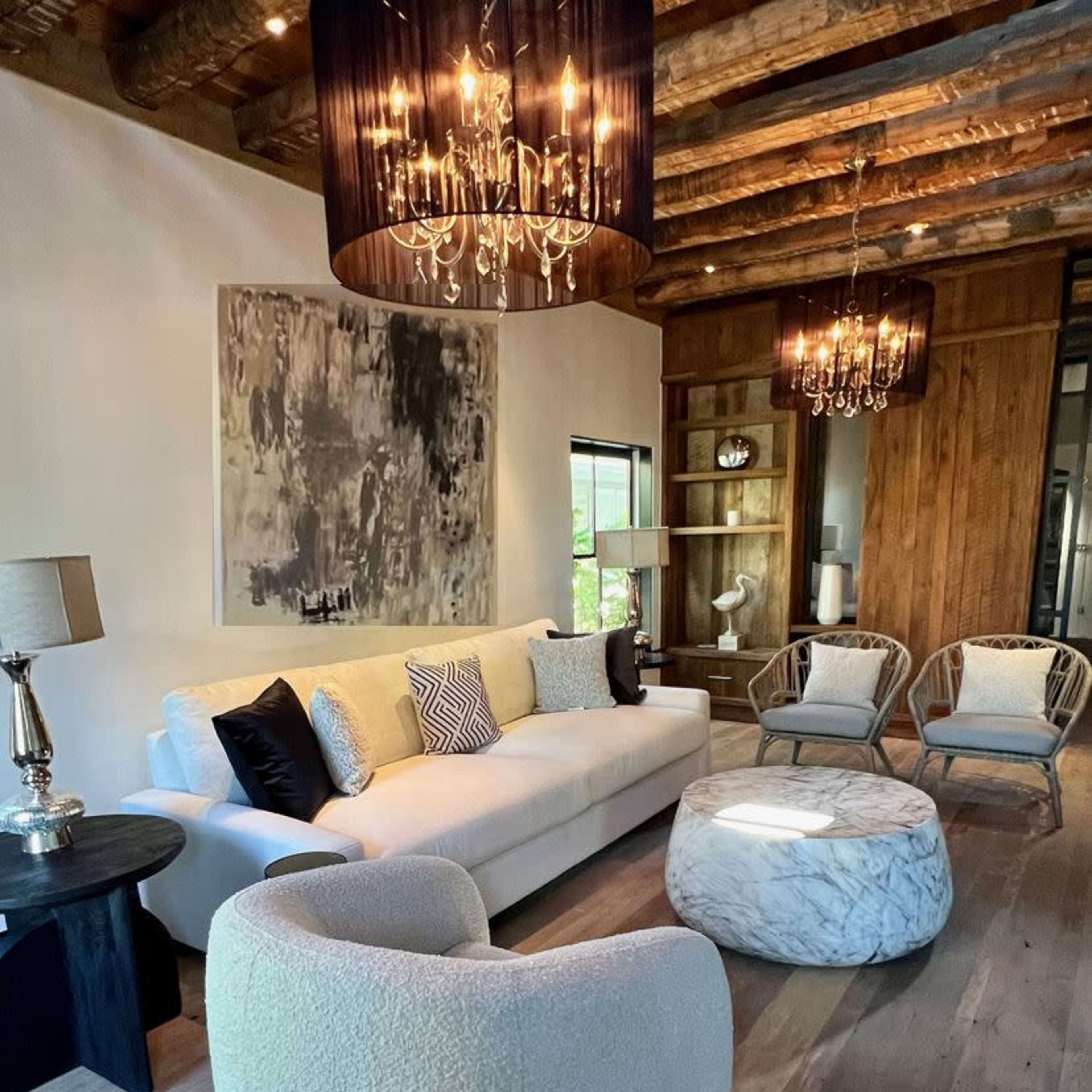 A modern living room with a white sofa, two accent chairs, a round marble coffee table, and several decorative elements under a ceiling with exposed beams and chandeliers.