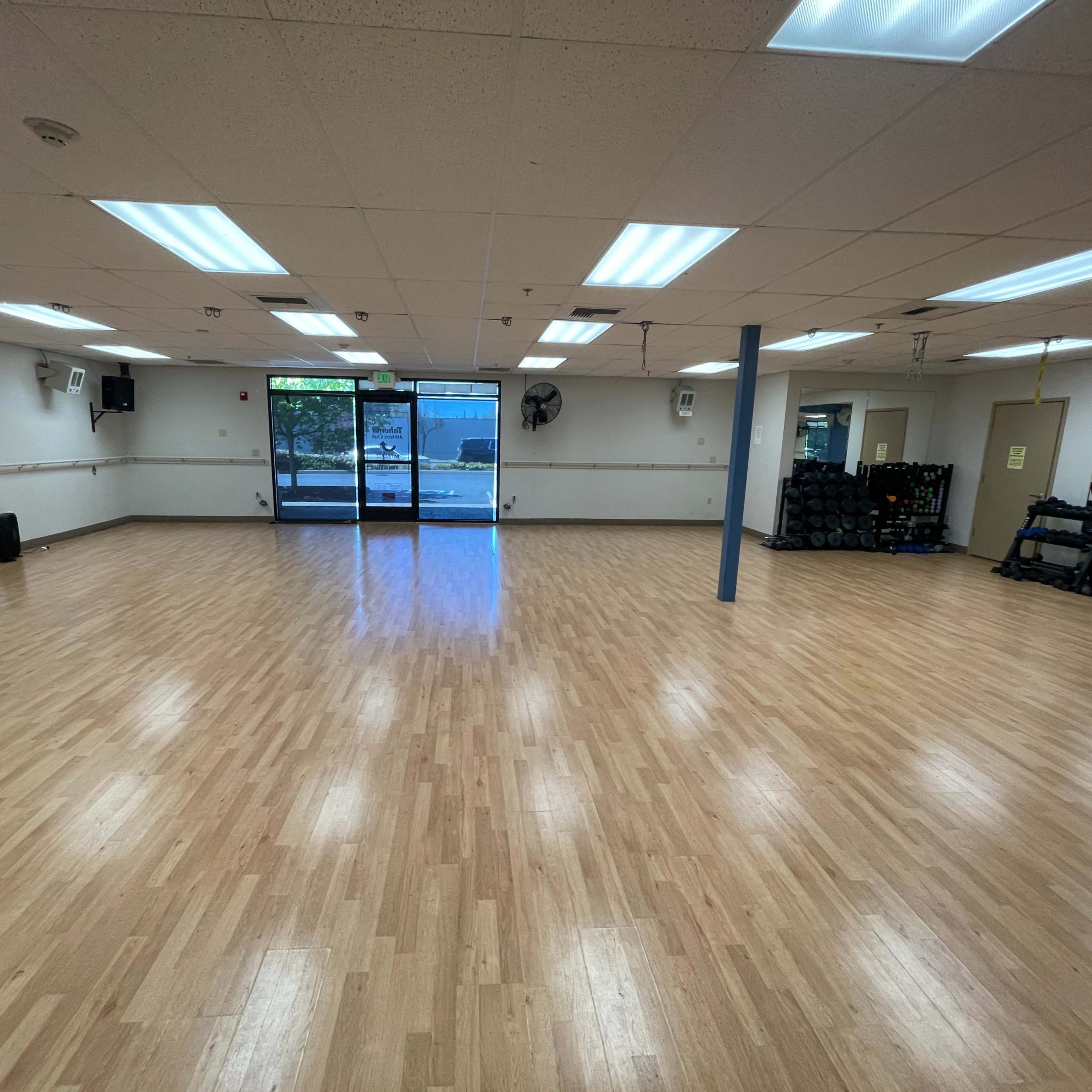 A spacious, well-lit fitness studio with wooden flooring and large windows at one end.