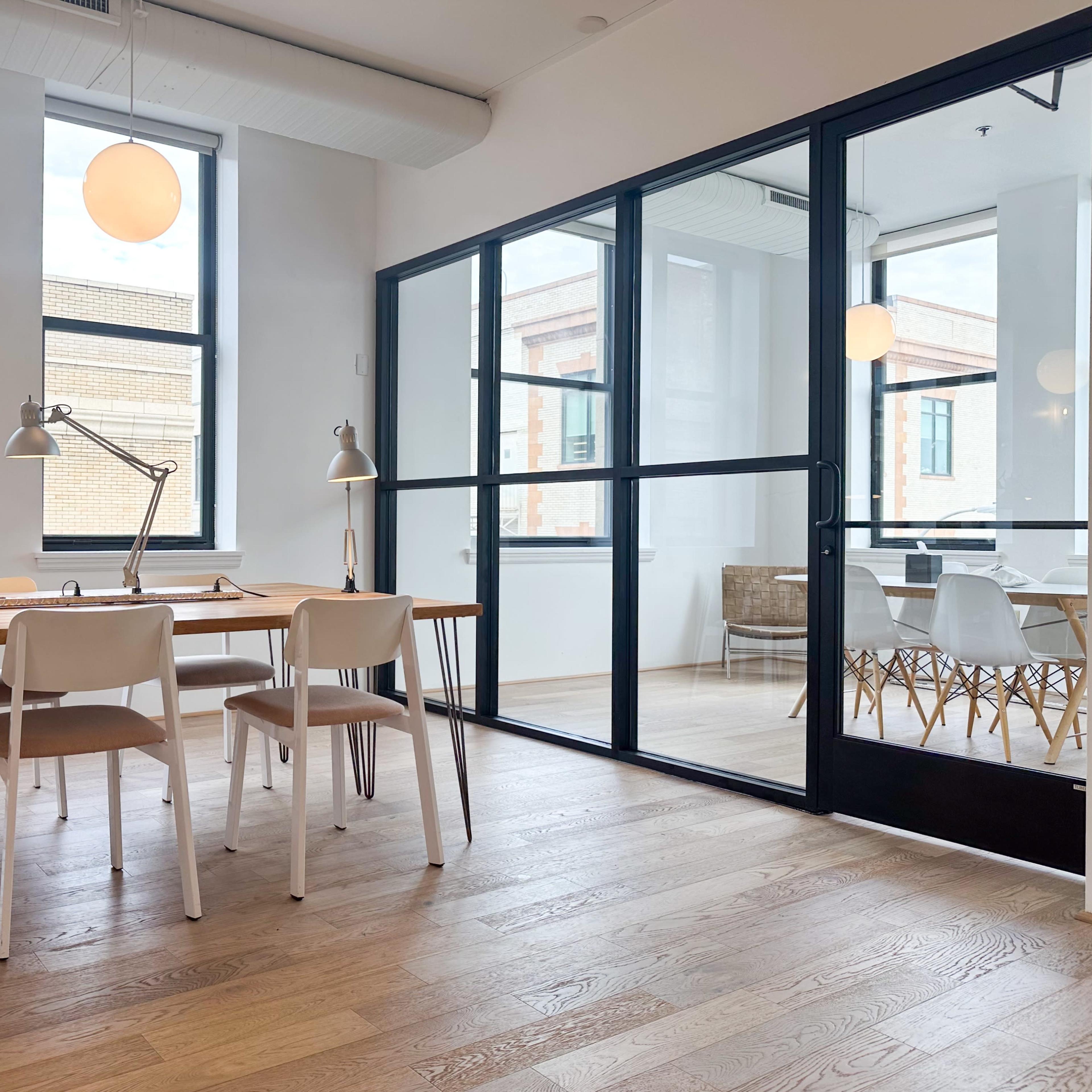 A modern office space with a wooden table and chairs on one side, and a glass-walled meeting room on the other side.