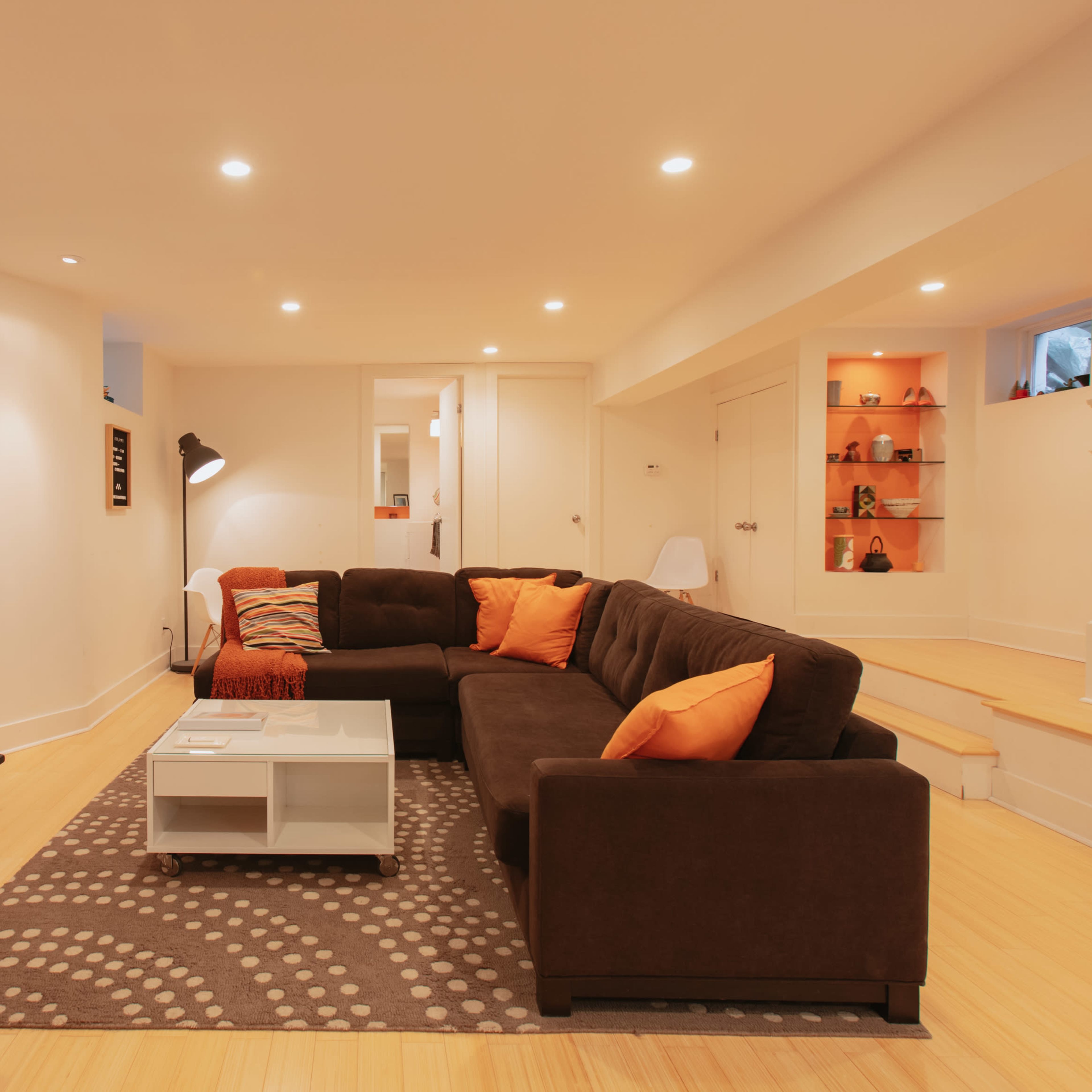 A well-lit basement living room featuring a dark sectional sofa with orange pillows, a white coffee table, a flat-screen TV, and a light-colored staircase.