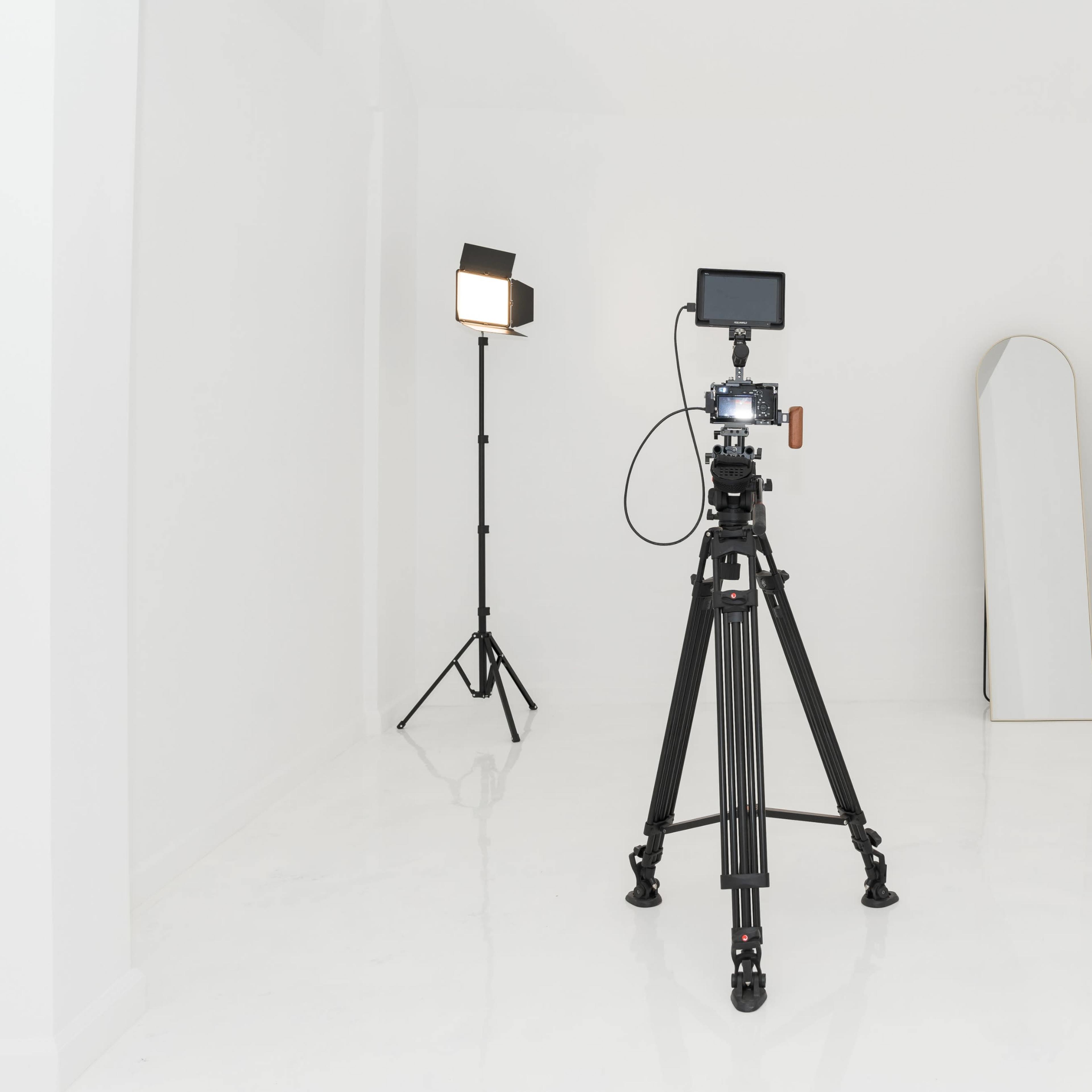 A camera on a tripod is positioned in an empty white room with a lighting setup and a mirror.