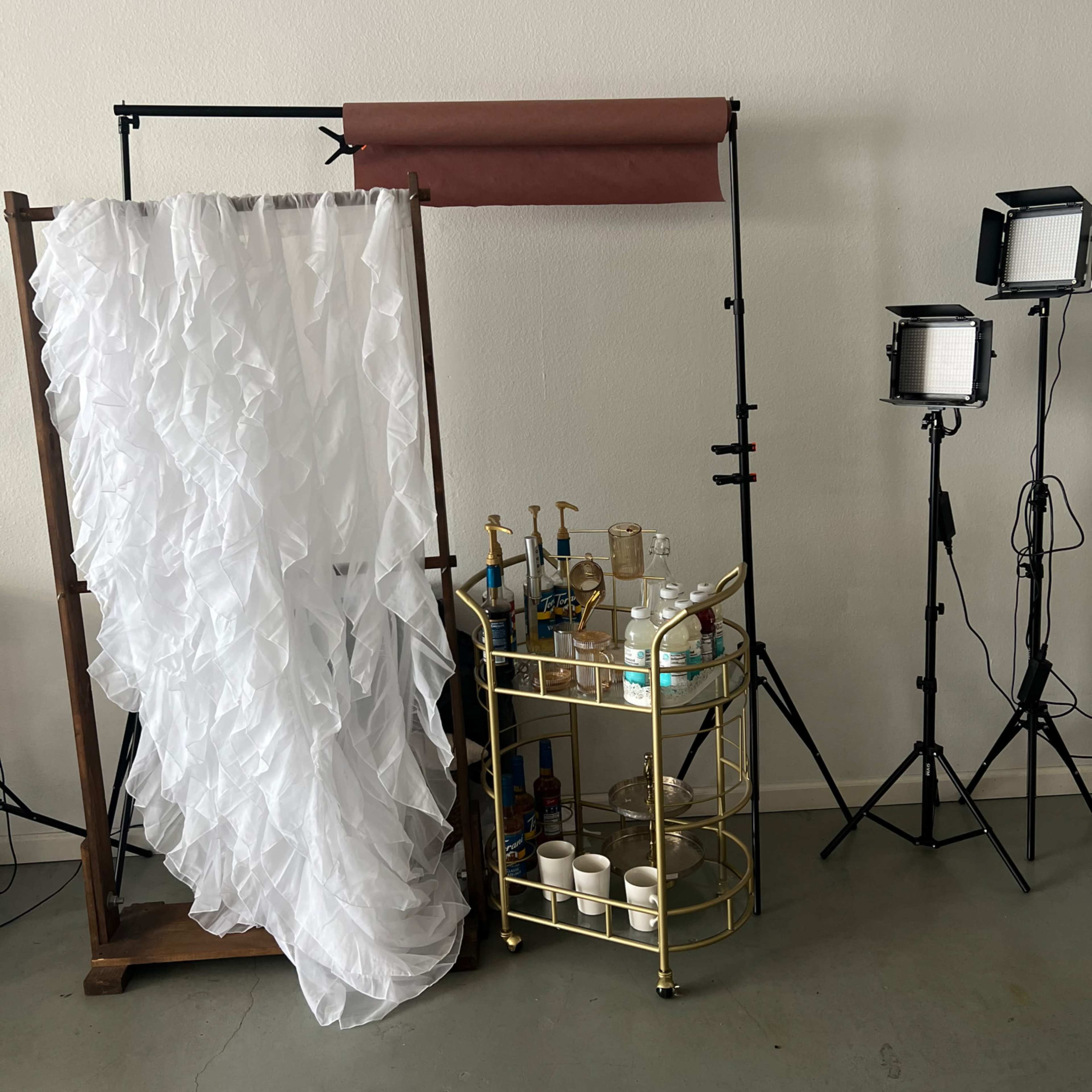 A white fabric drapes from a wooden frame beside a golden cart filled with bottles and containers, illuminated by two light stands.