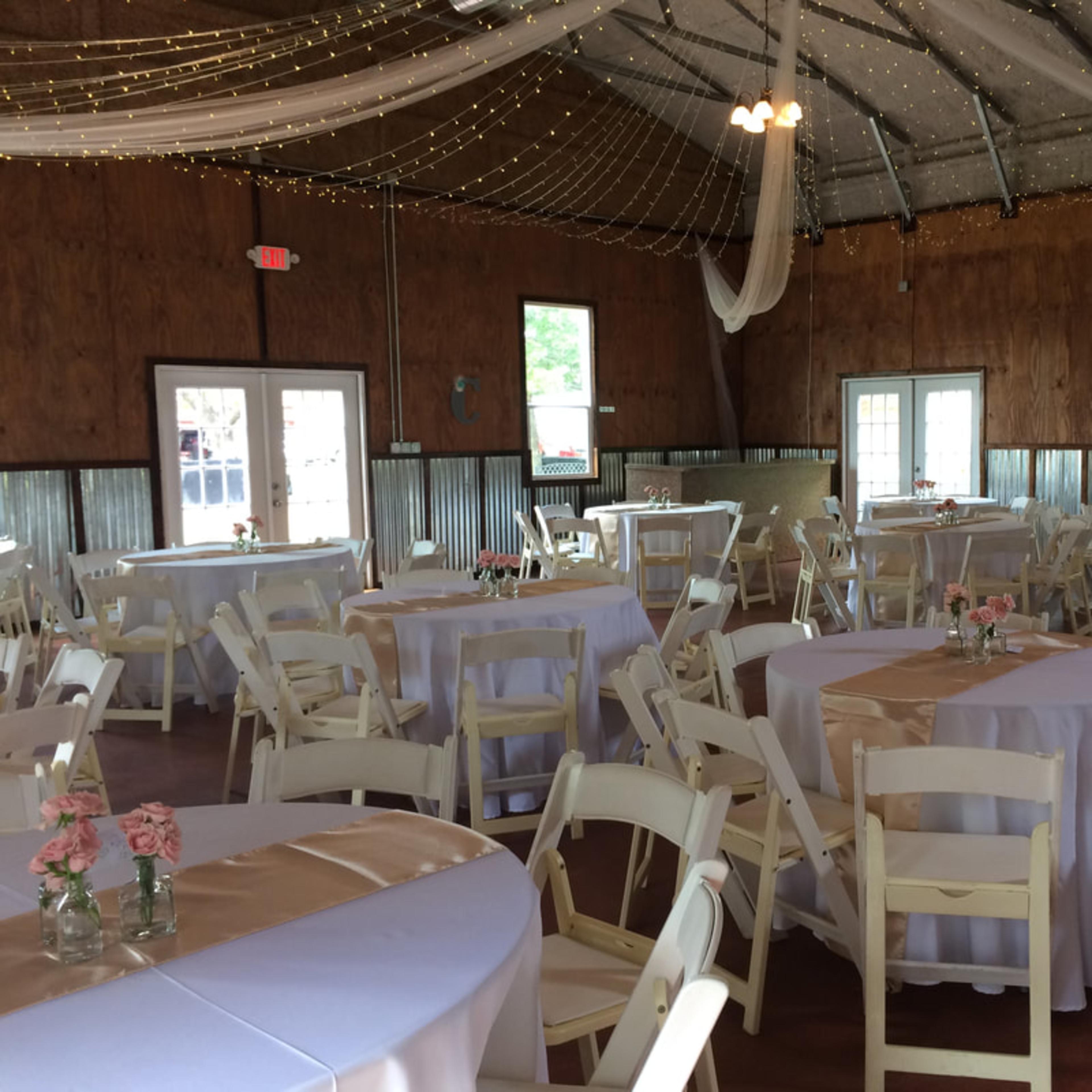 A spacious event venue with round tables covered in white tablecloths and pink decorative elements, surrounded by a wooden interior and string lights overhead.