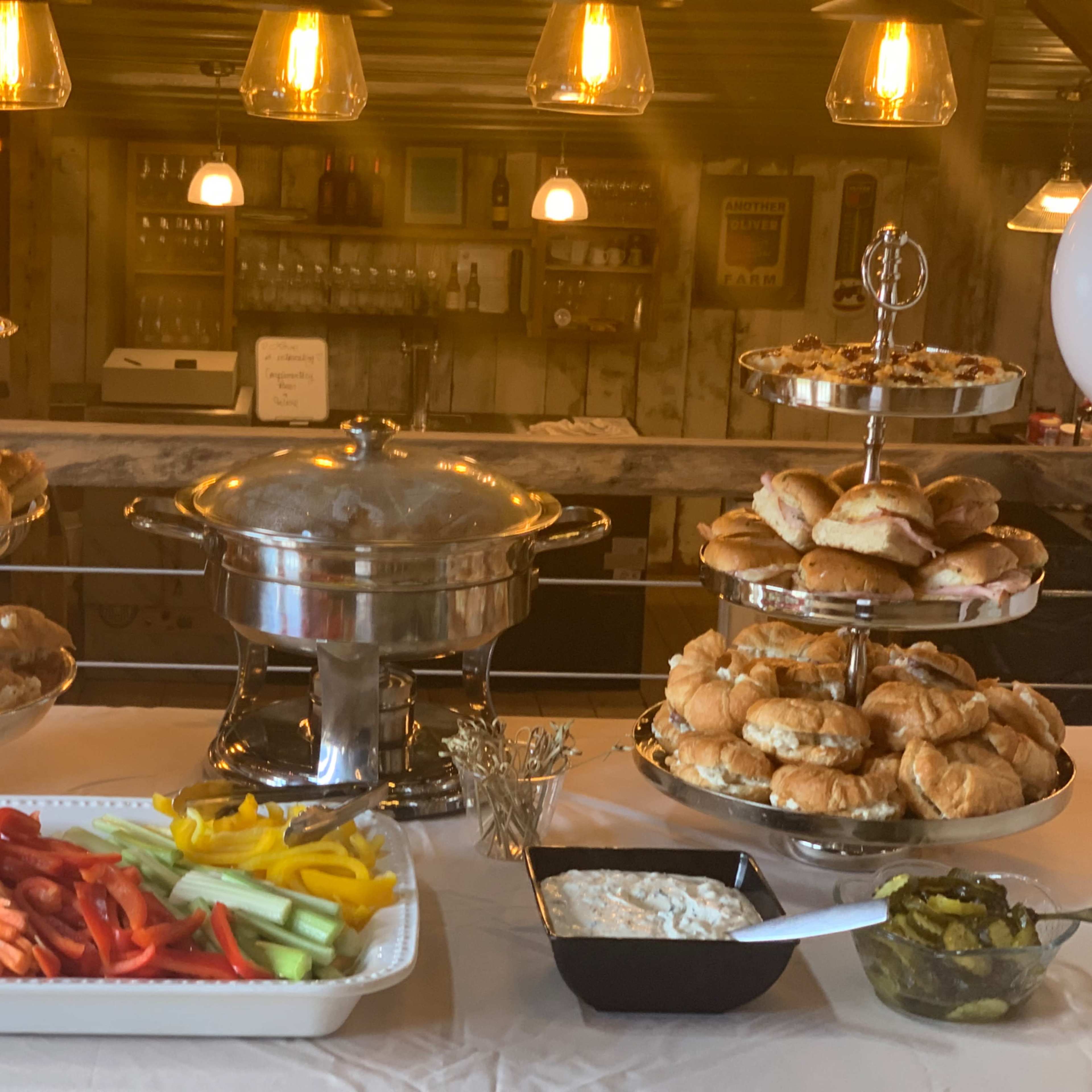 A buffet setup features two tiers of assorted sandwiches and pastries, a chafing dish with hot food, and a platter of fresh vegetables and dip.