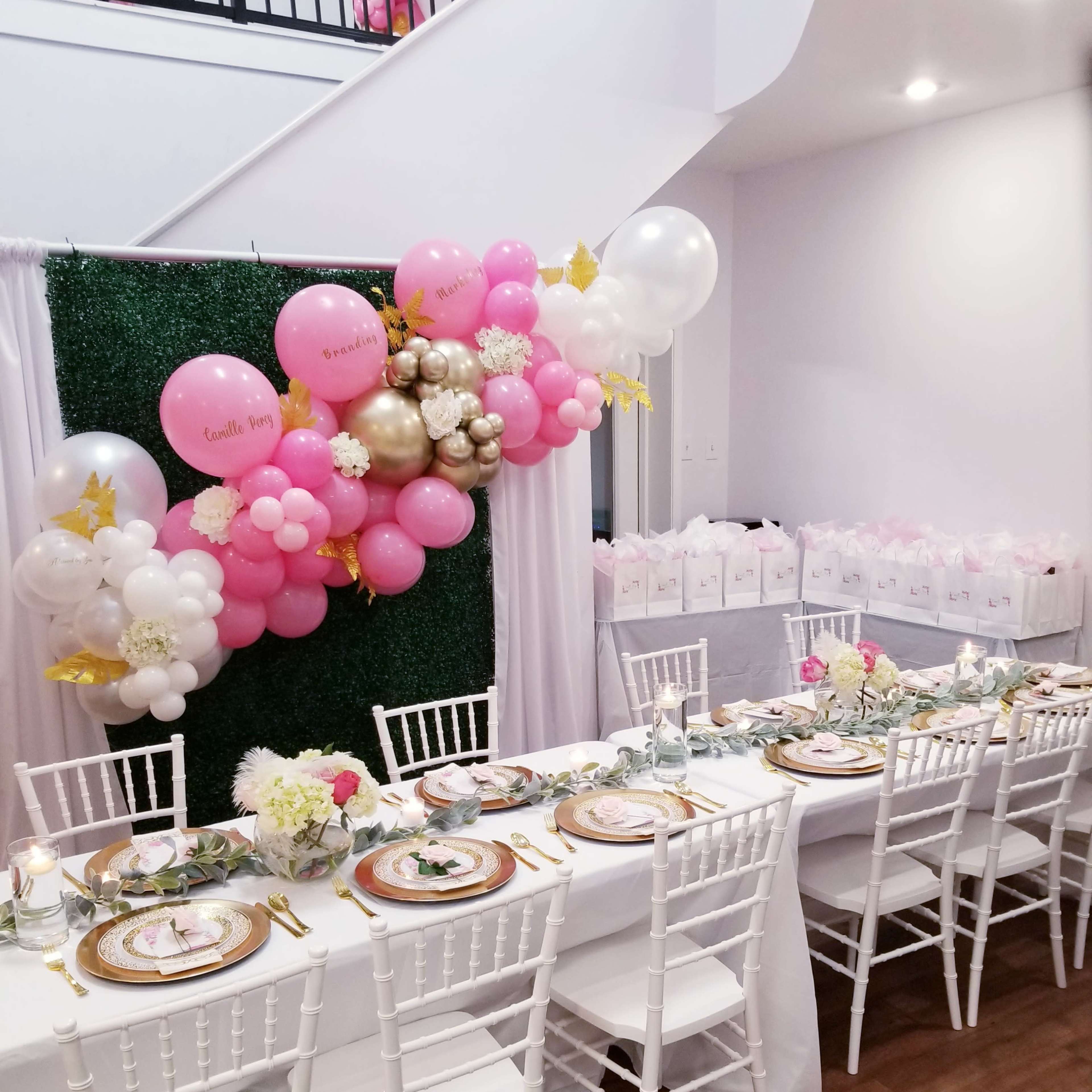 A long table is set with floral arrangements and gold plates, surrounded by white chairs, against a backdrop of a decorated wall with balloons and gift bags on a nearby surface.
