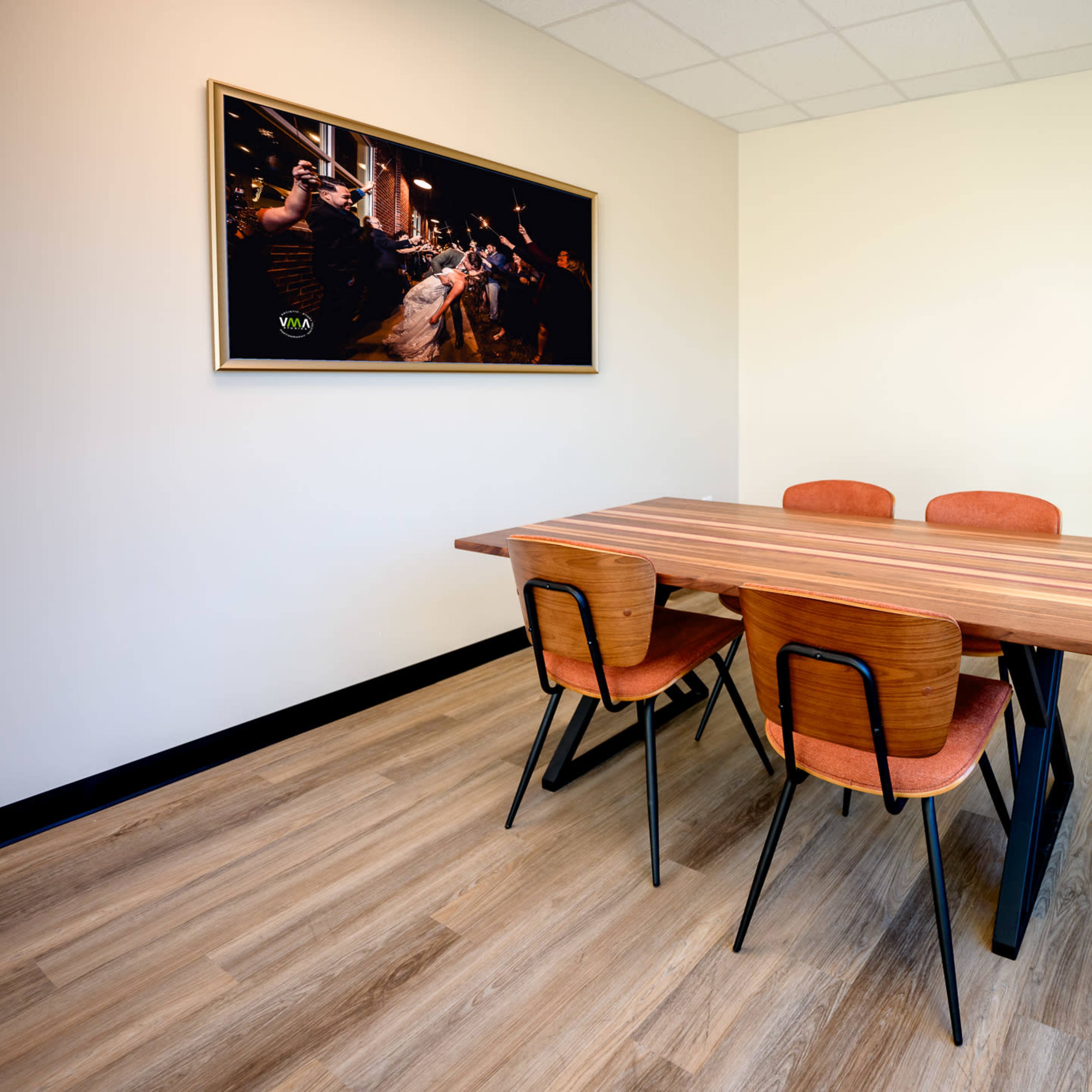 A wooden table with four chairs is positioned in a sparse room featuring light-colored walls and a large framed photograph on one wall.