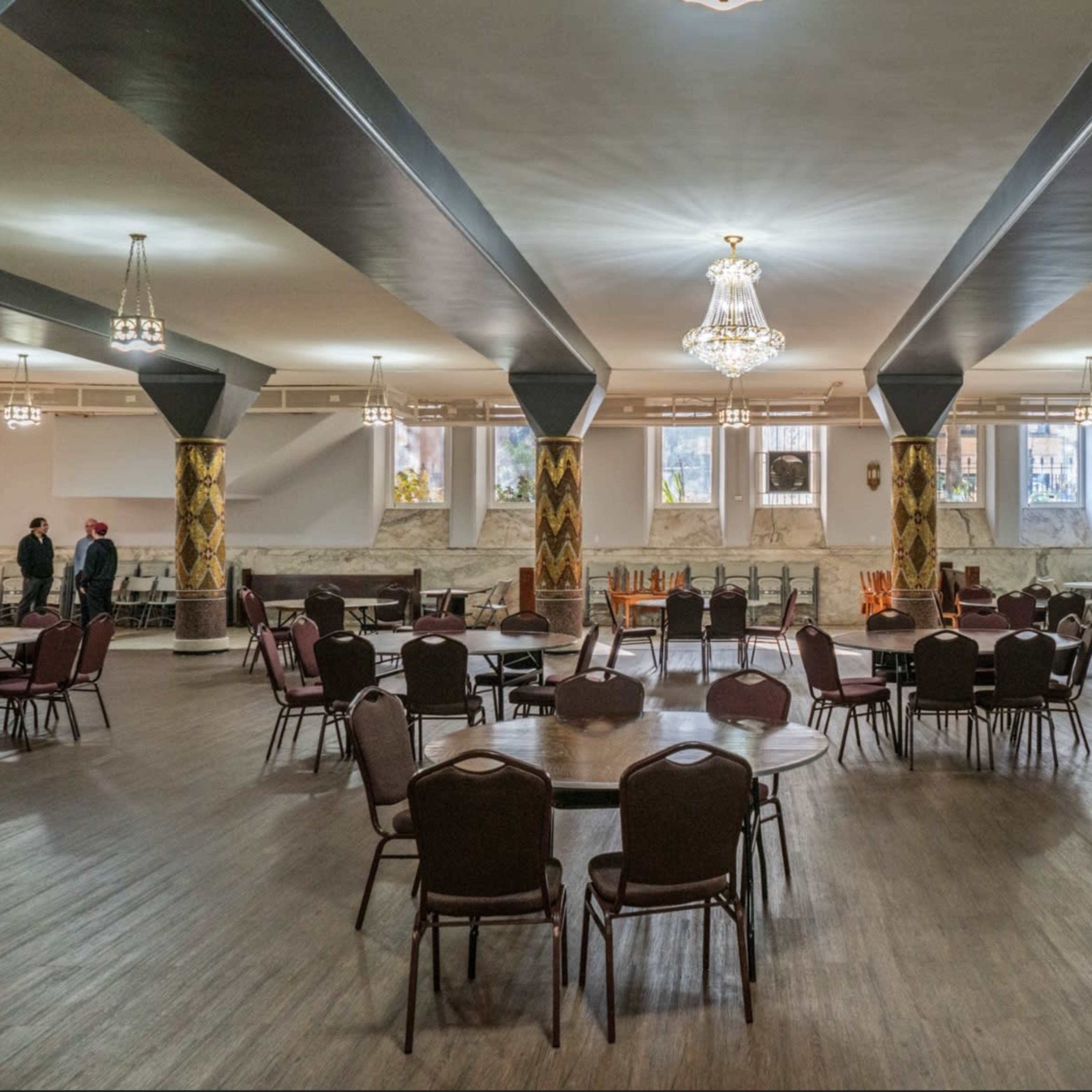 A spacious dining hall featuring several round tables surrounded by chairs, with decorative pillars and chandeliers overhead.