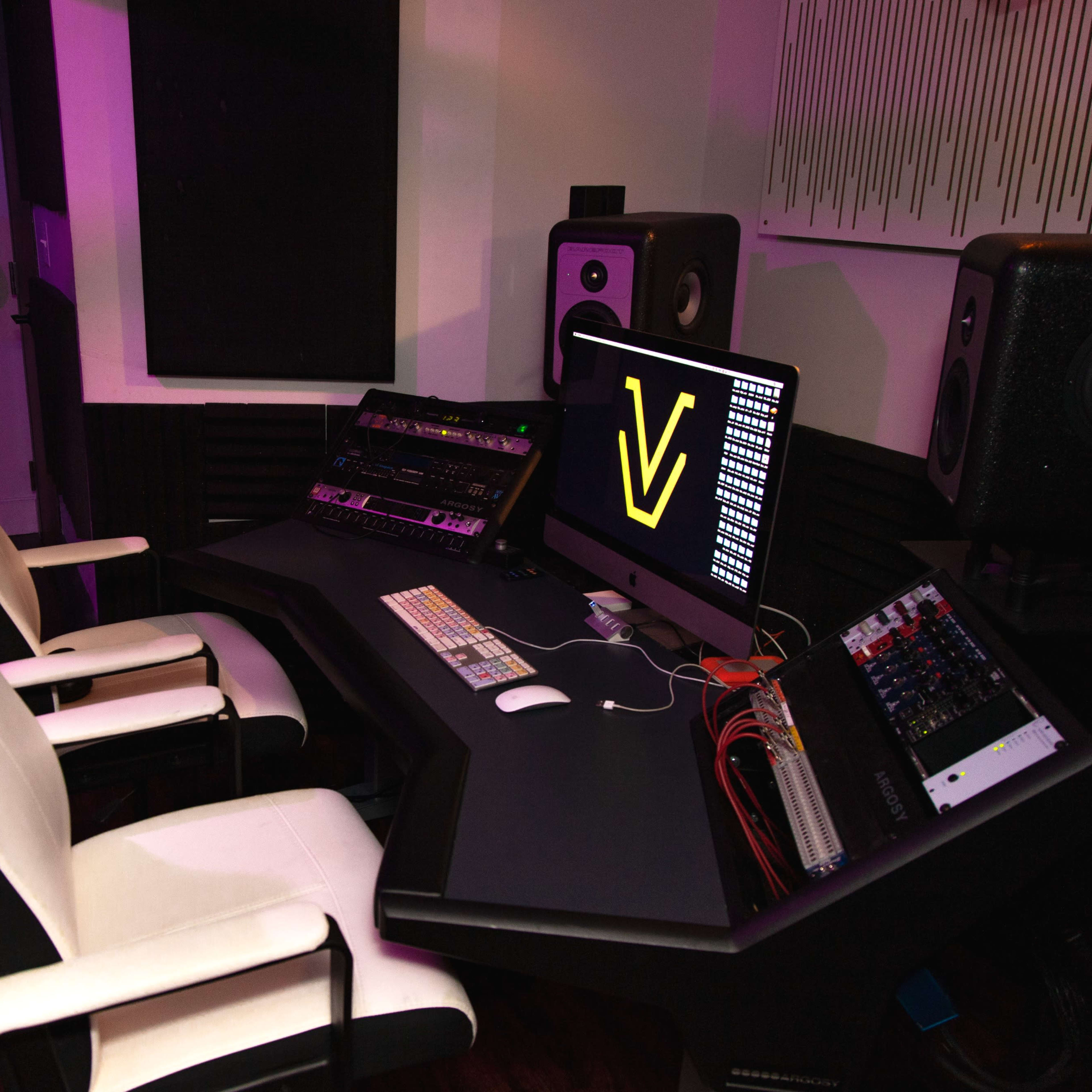 A modern music studio with a sleek black desk, two ergonomic white chairs, computer equipment, and two audio speakers on either side.
