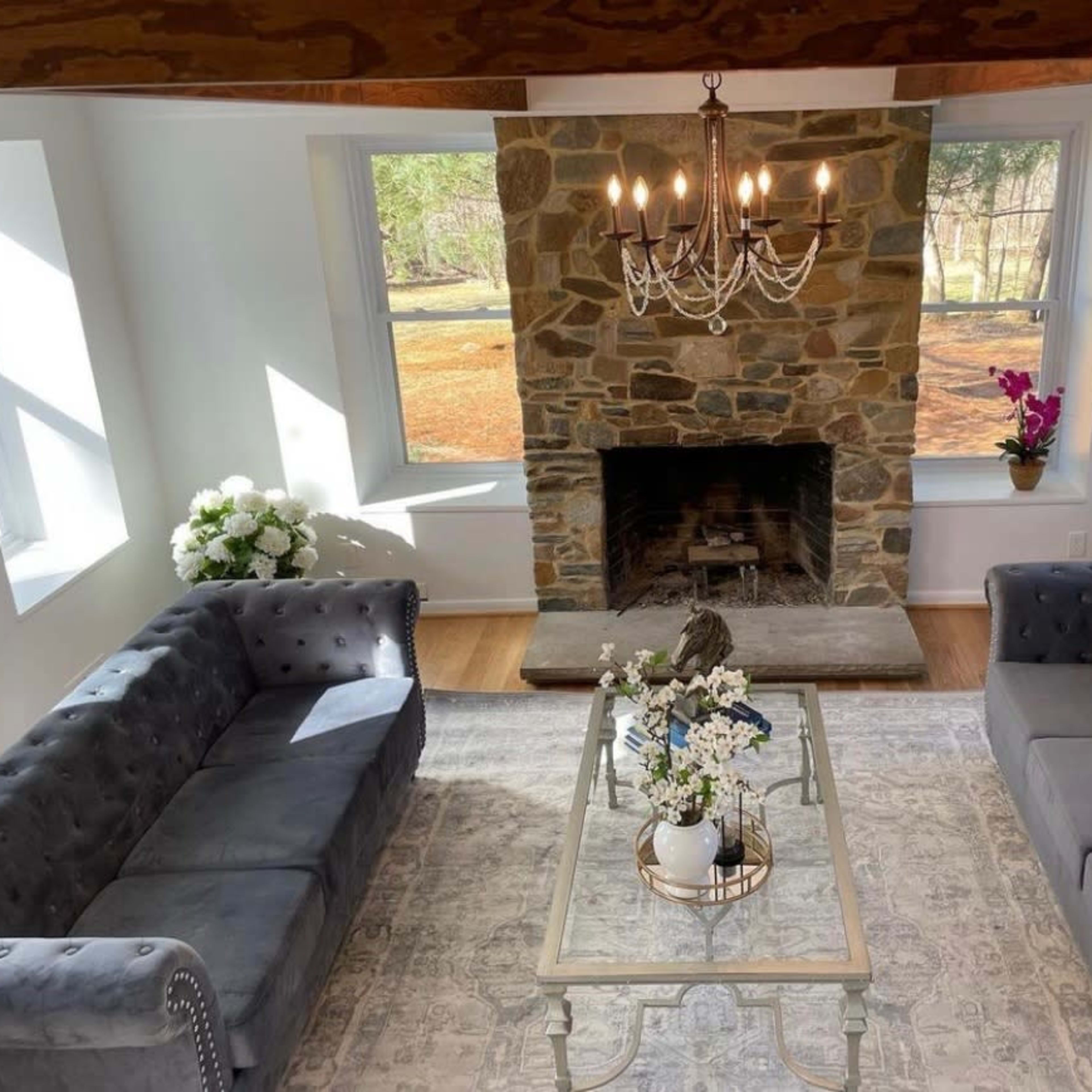 A bright living room featuring two gray tufted sofas, a coffee table with floral arrangements, and a stone fireplace beneath a chandelier.