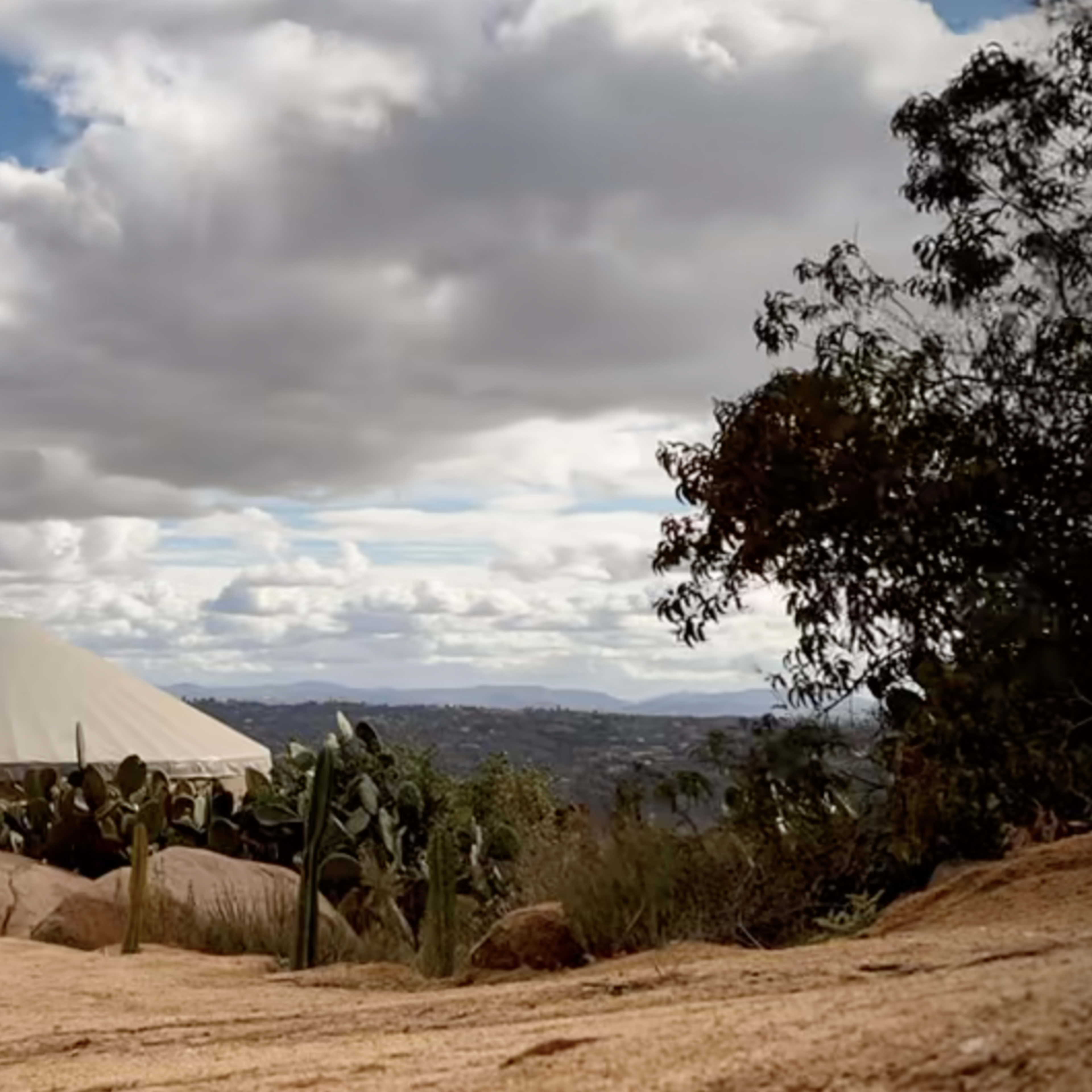 A yurt stands on a rocky path surrounded by cacti and a view of distant hills under a cloudy sky.