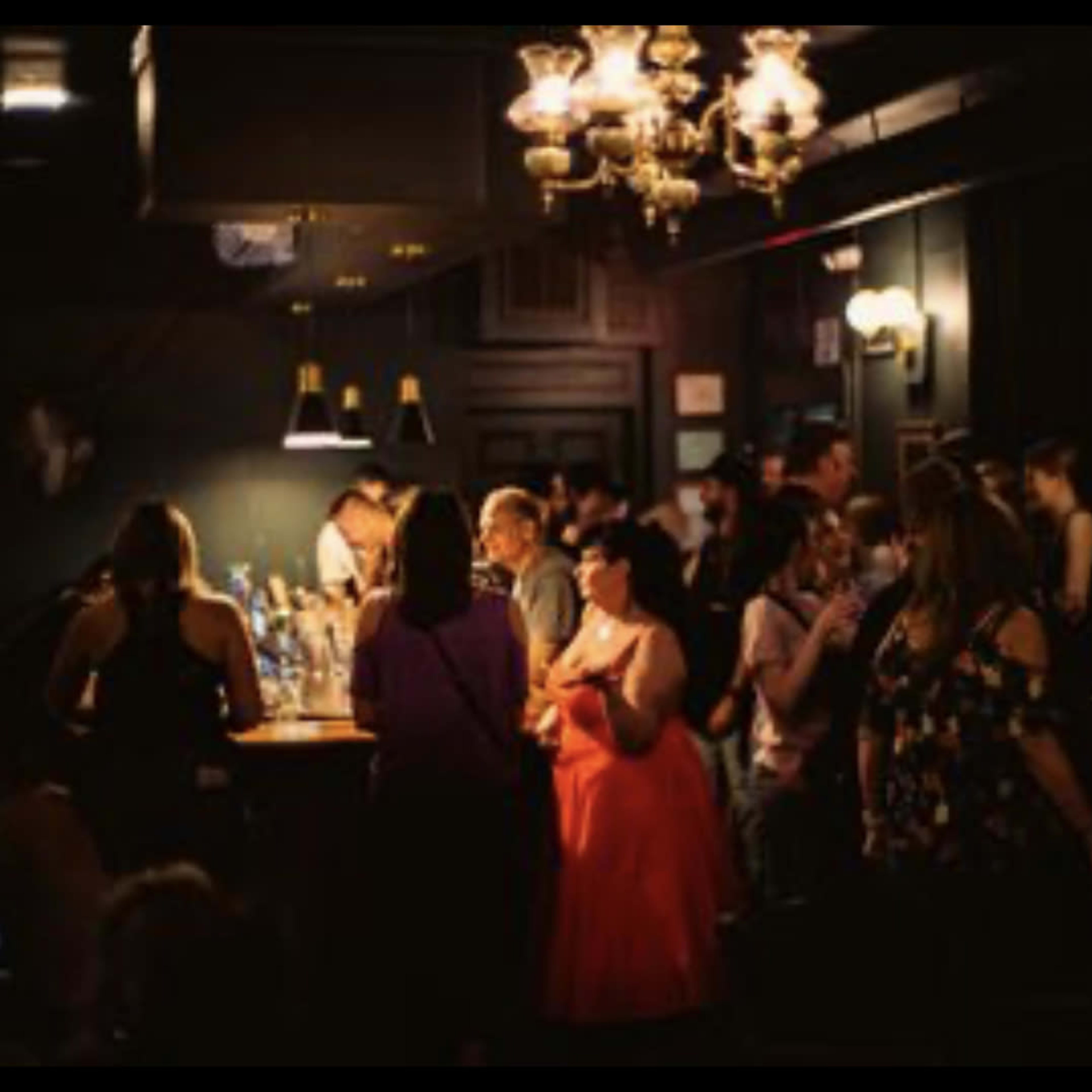 A crowded bar scene features patrons gathering around a well-lit bar while others socialize in a dimly lit environment.