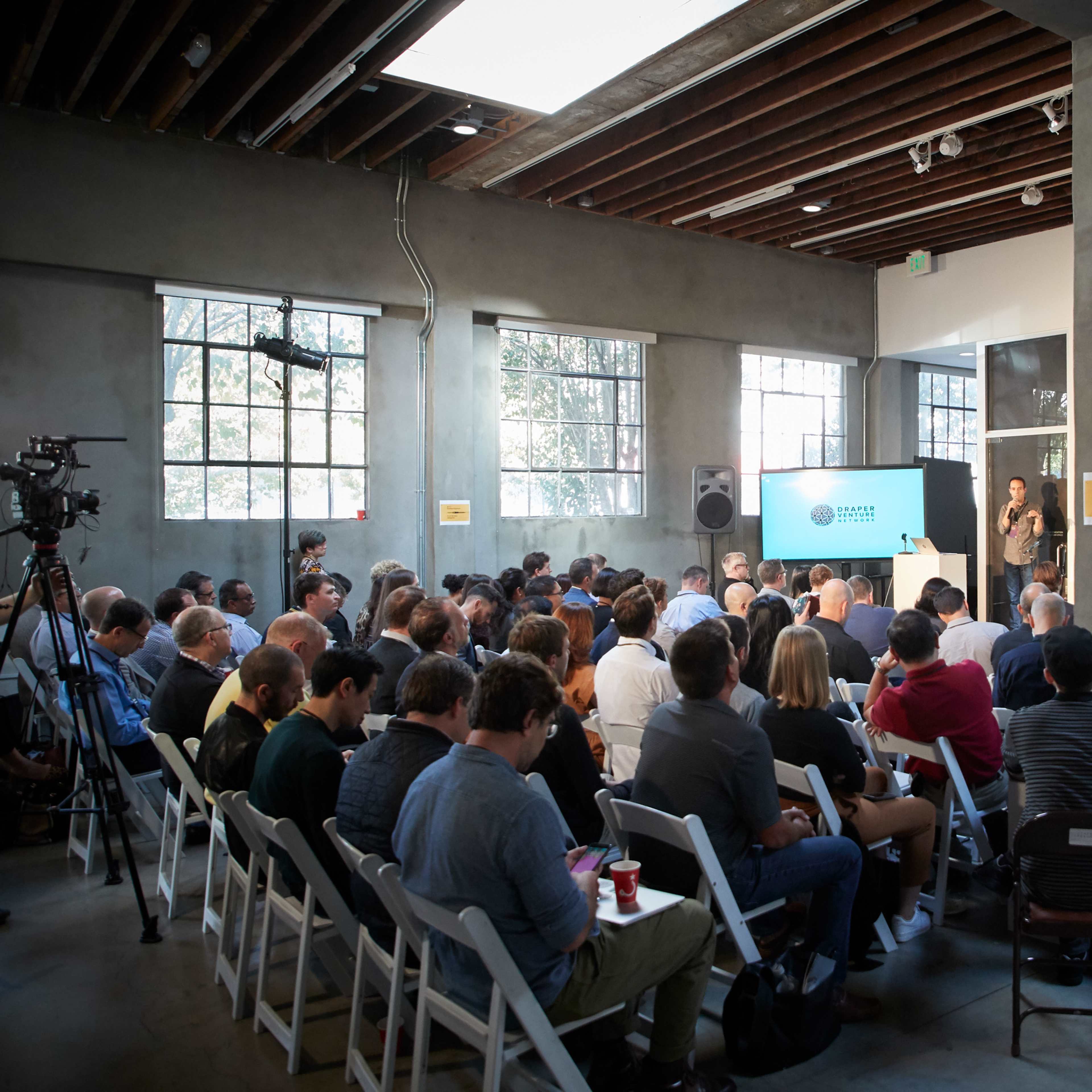 A group of people sits in rows facing a presentation screen in a spacious, well-lit indoor venue.
