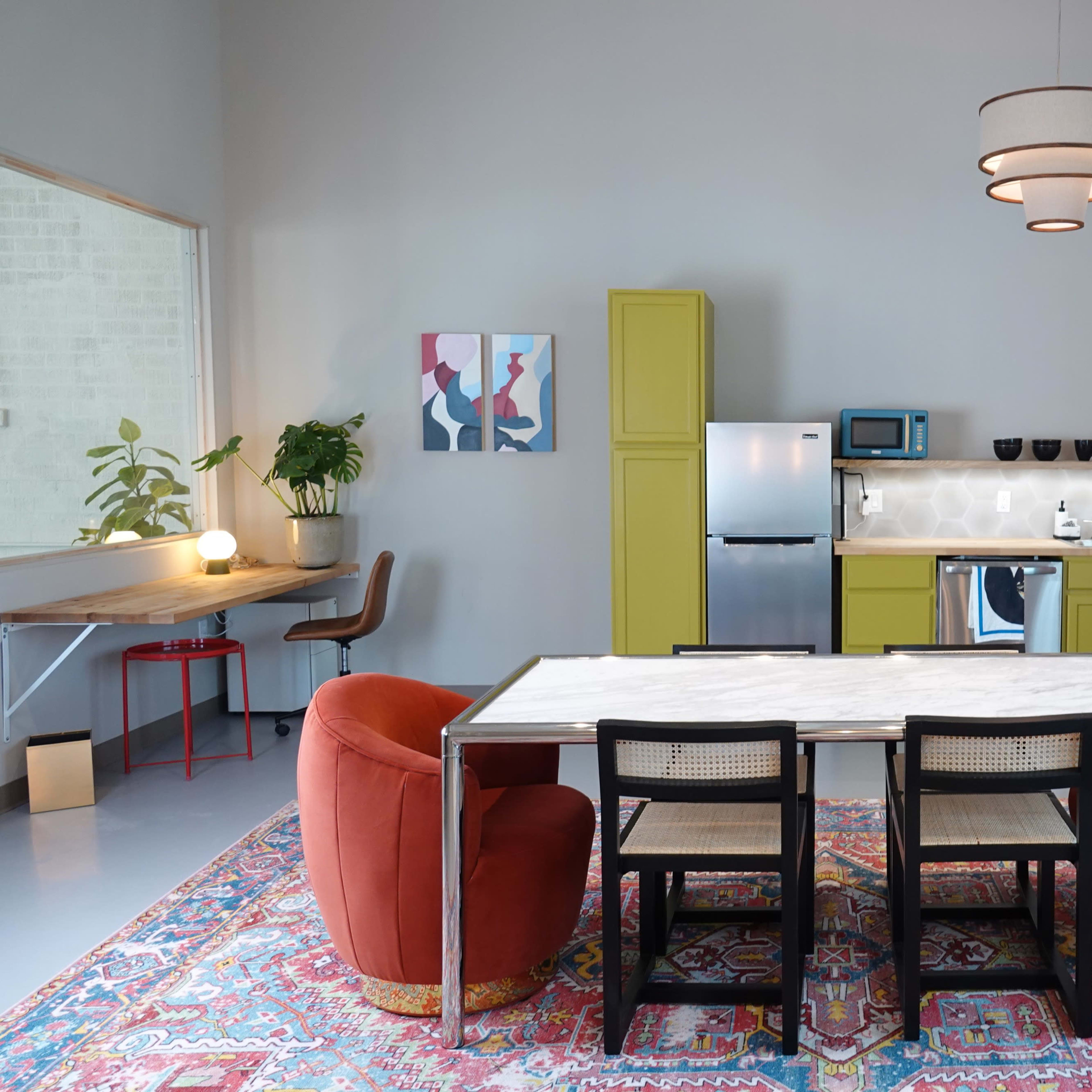 A modern kitchen and dining area features a marble-top table surrounded by black chairs, flanked by two red armchairs, with olive green cabinets and colorful artwork on the walls.