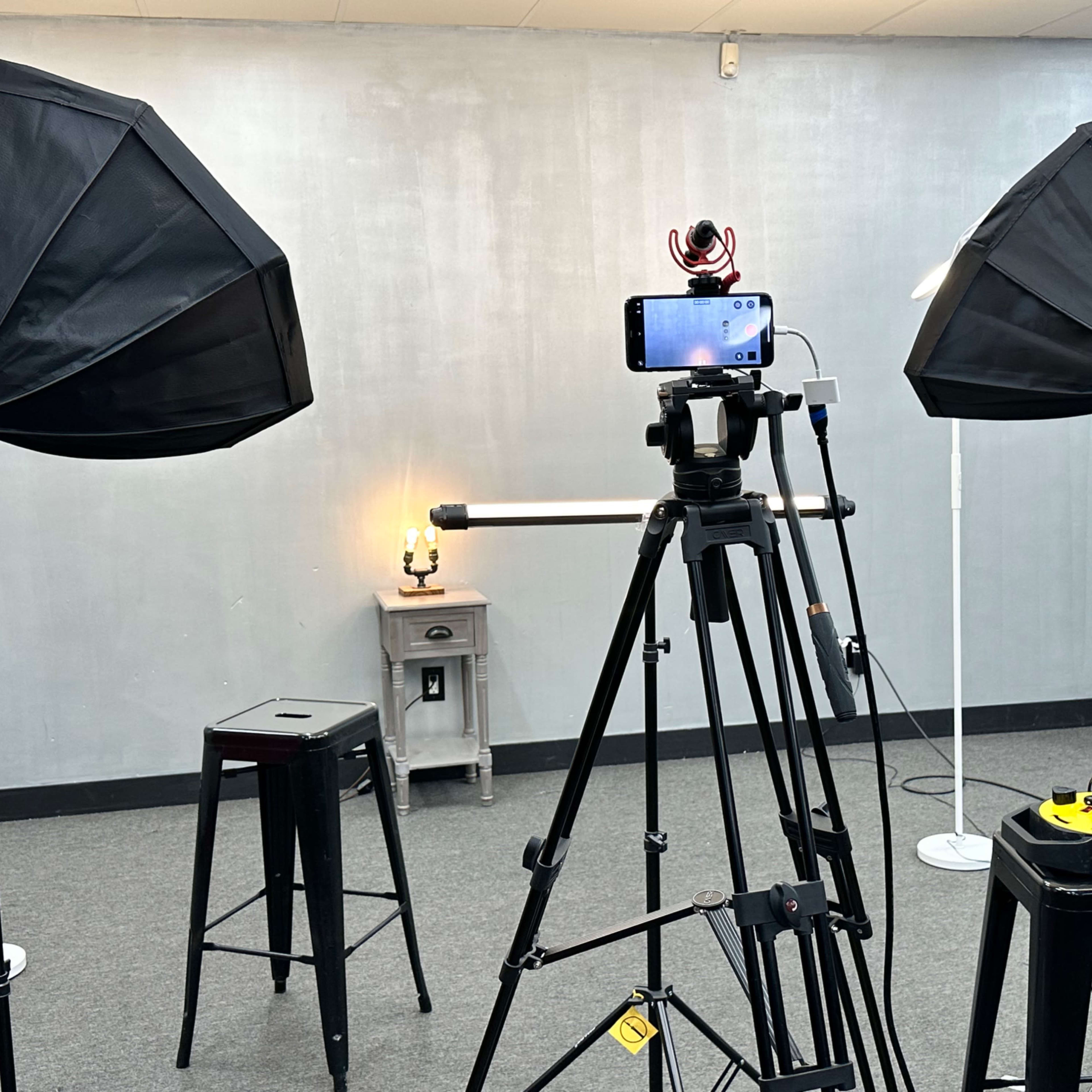 A photography setup with two softbox lights, a smartphone mounted on a tripod, a stool, and a small table with a lamp in a room with gray walls.