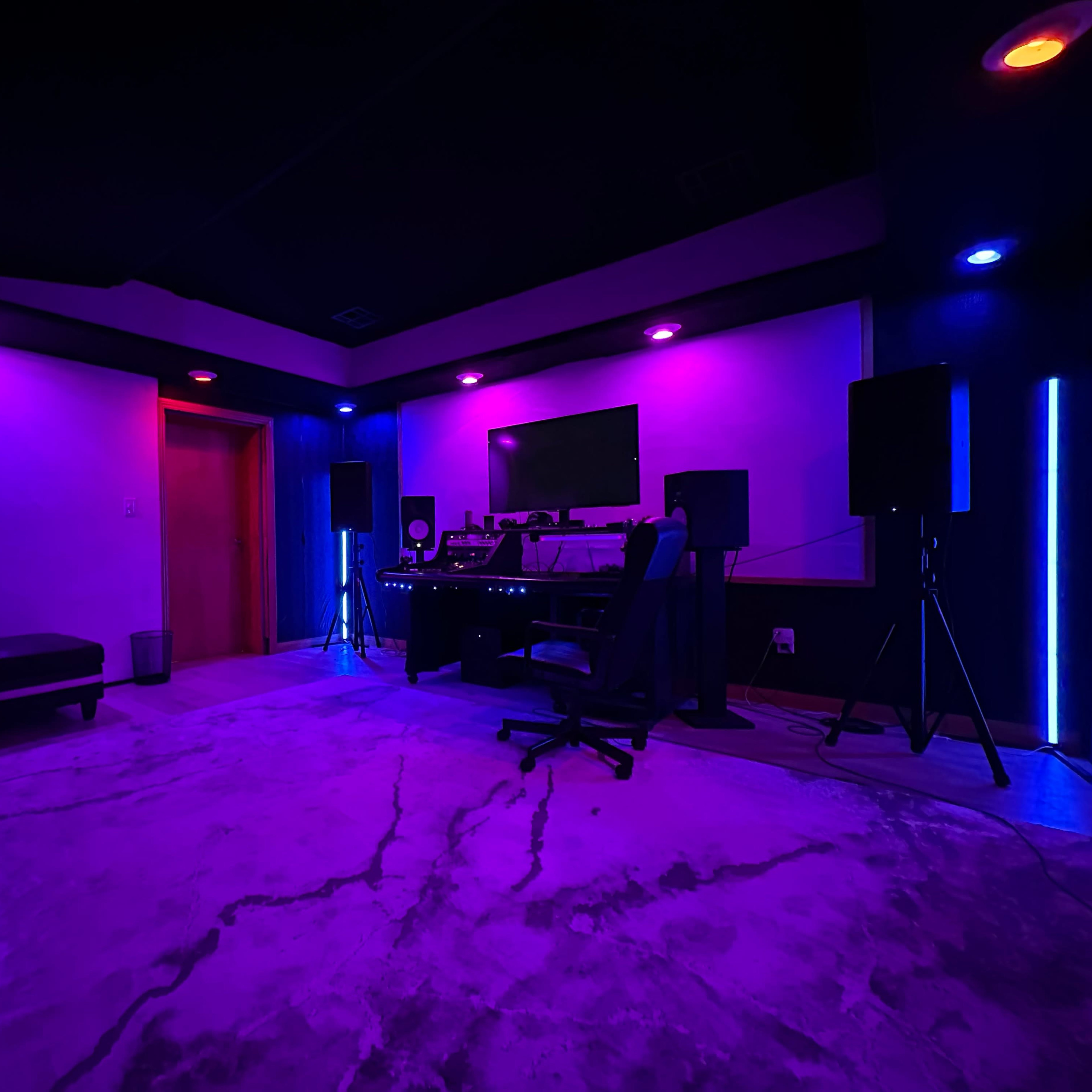 A modern recording studio with purple and blue lighting, featuring a desk with audio equipment, a chair, and a comfortable couch.