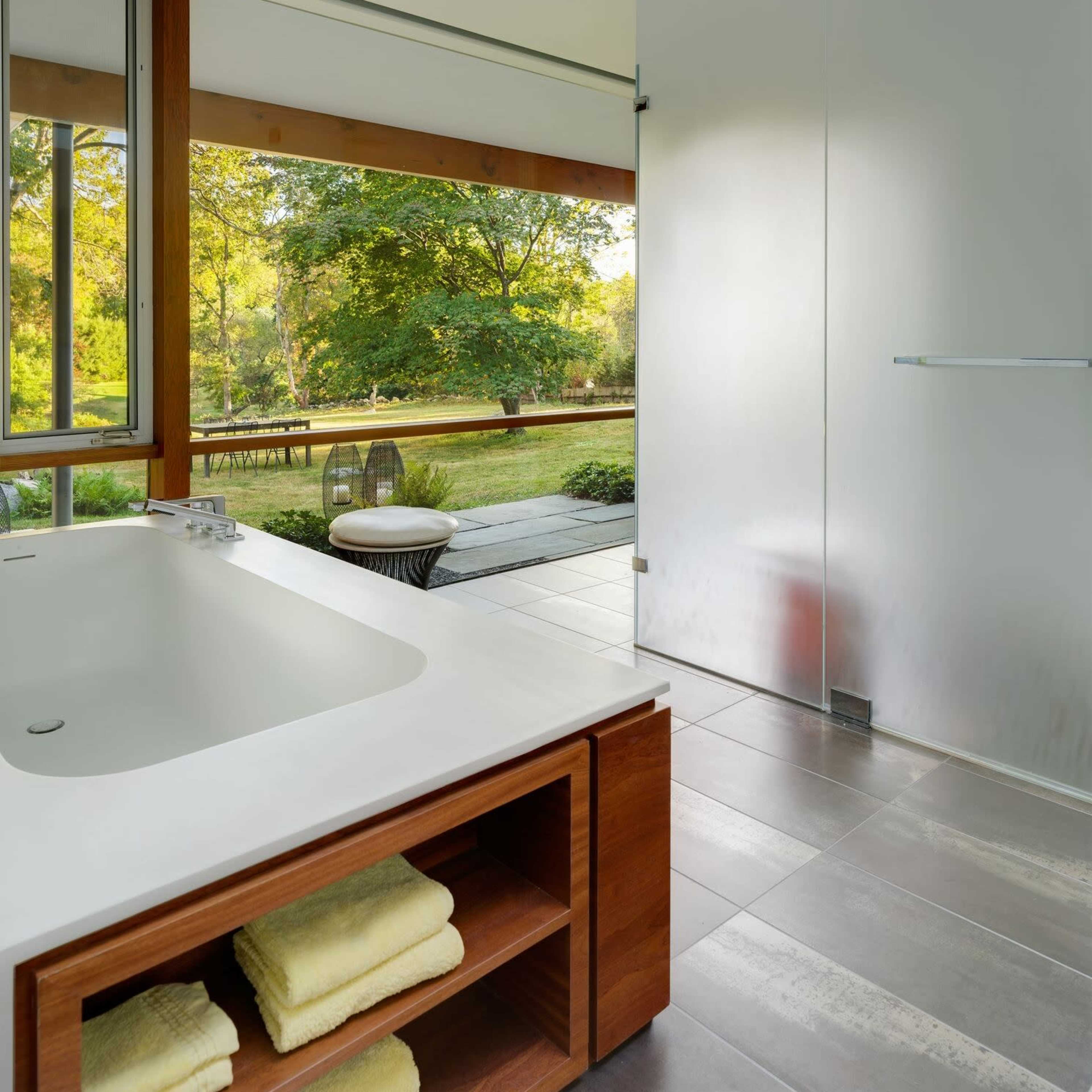 A modern bathroom features a large bathtub and a frosted glass shower door, opening to a view of a grassy outdoor area.
