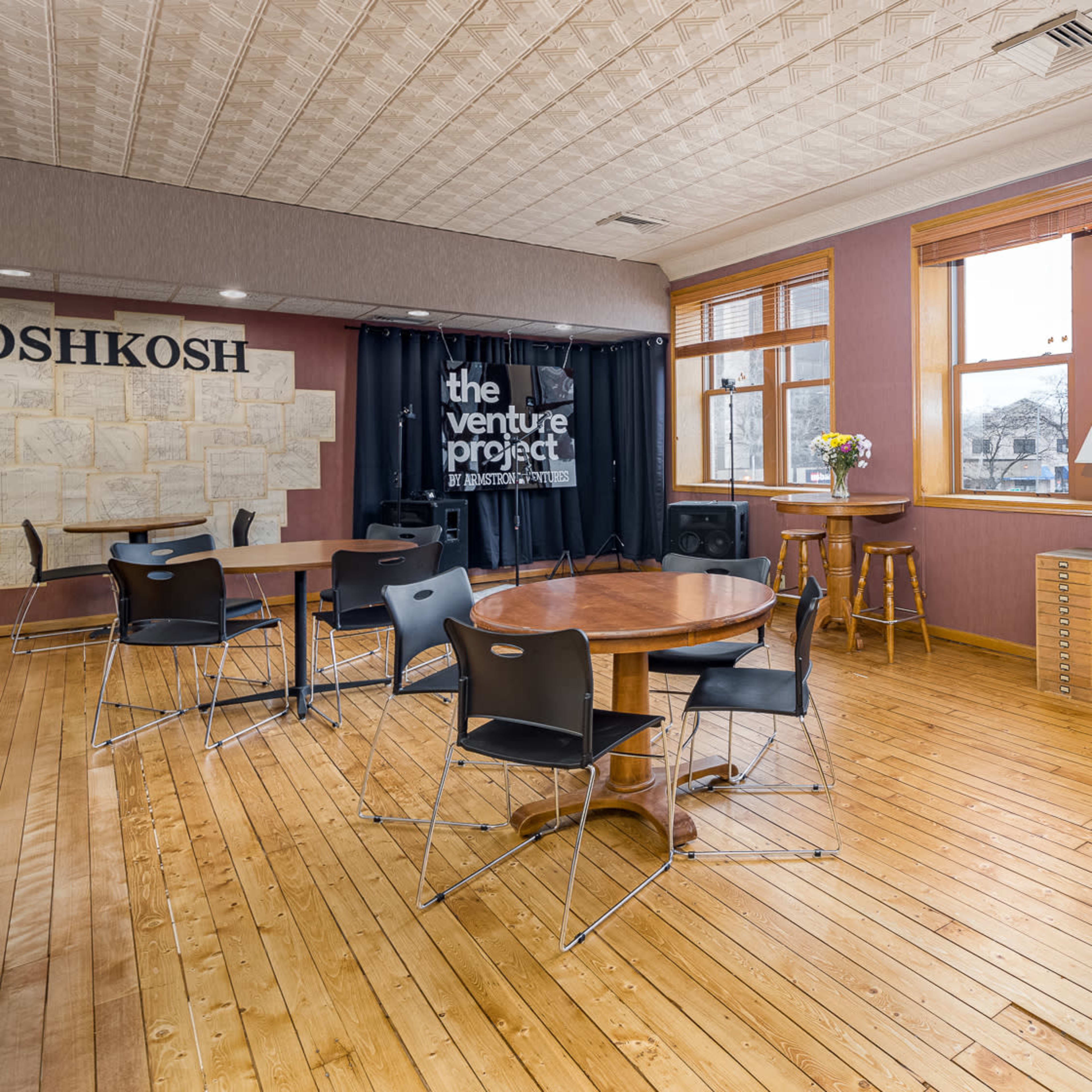 The room features wooden floors, large windows, a sofa, a round table with chairs, and a wall displaying a sign for "the venture project."