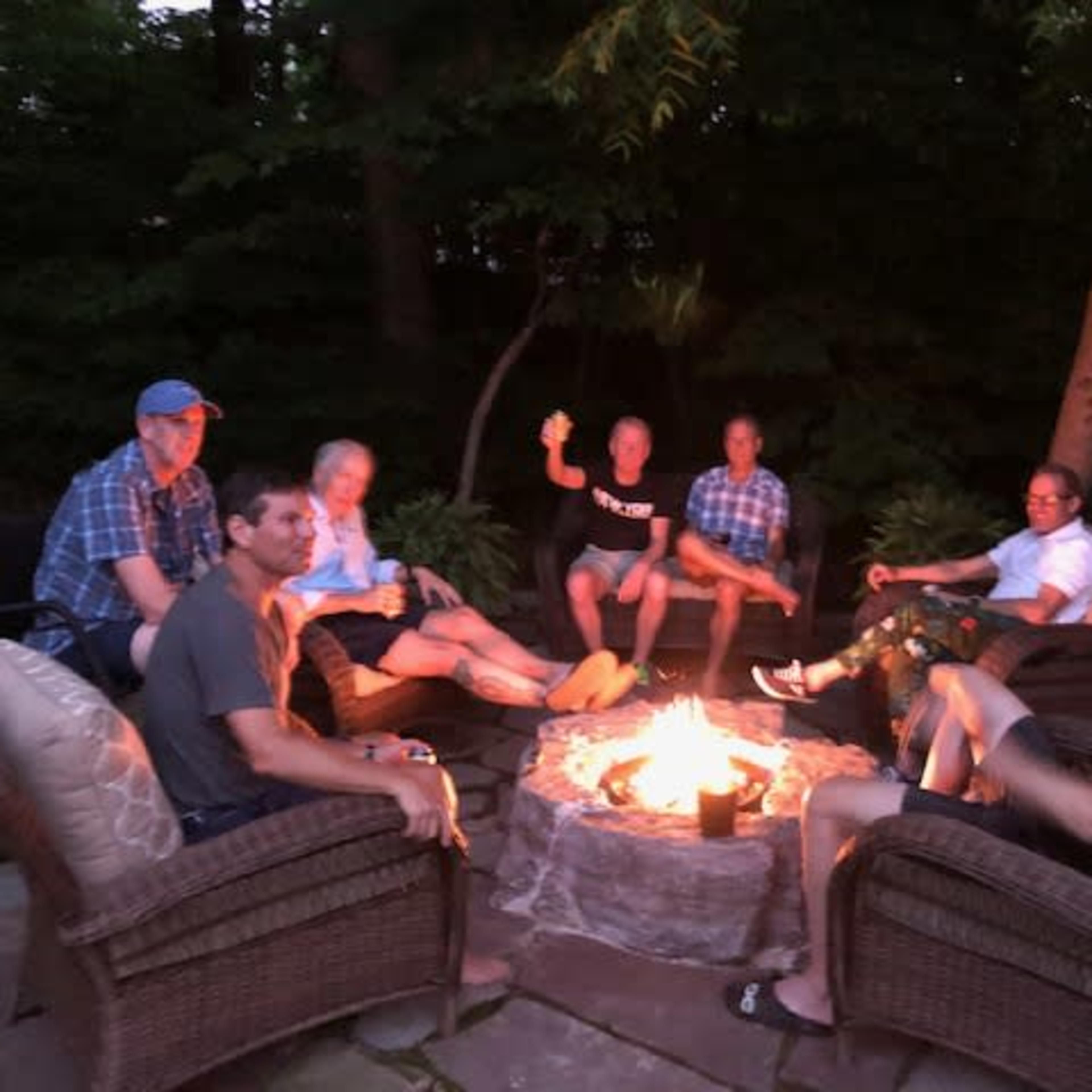 A group of seven men sits around a fire pit in a backyard, engaged in conversation as the fire burns brightly.