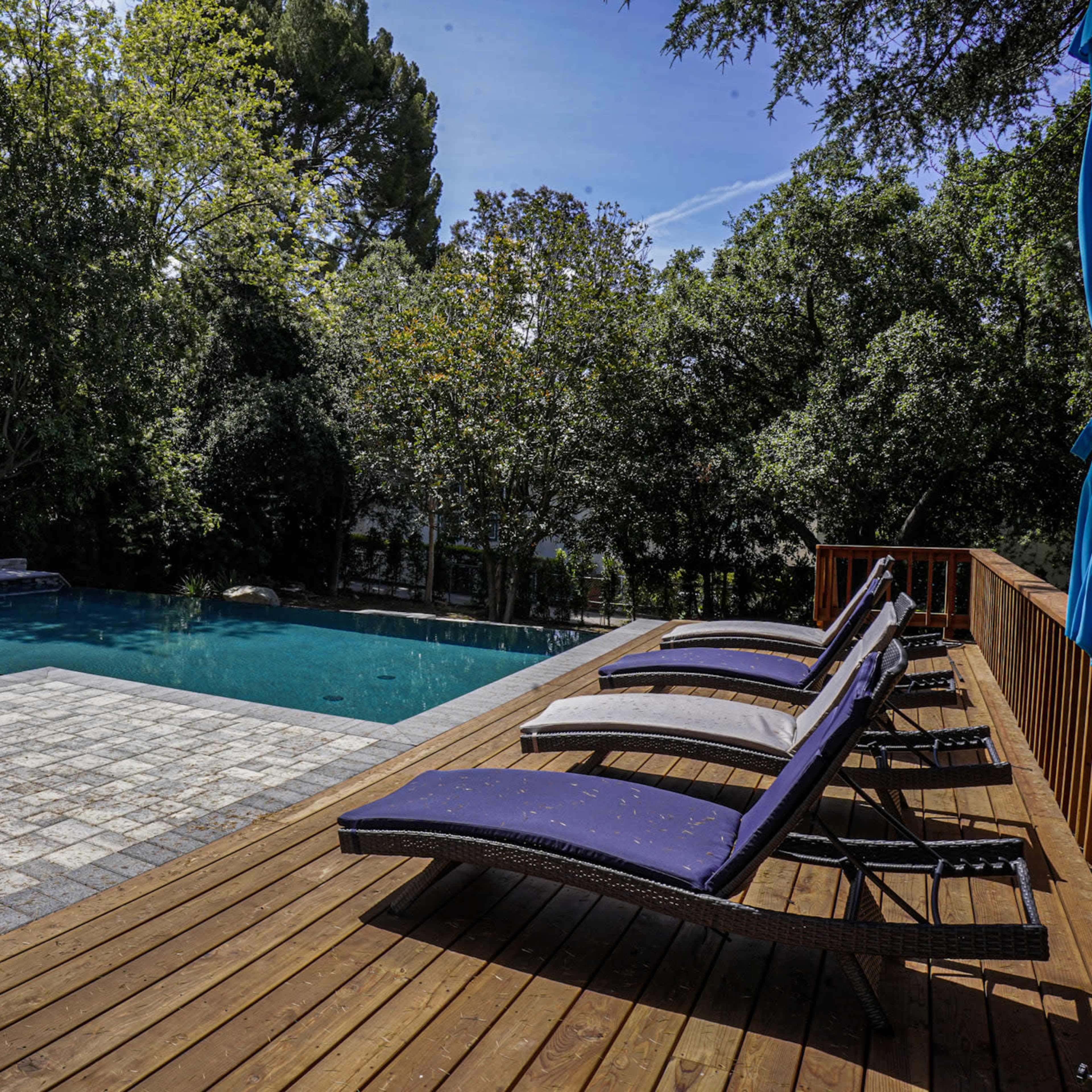 A rectangular swimming pool surrounded by wooden decking and lounge chairs, with lush green trees in the background.