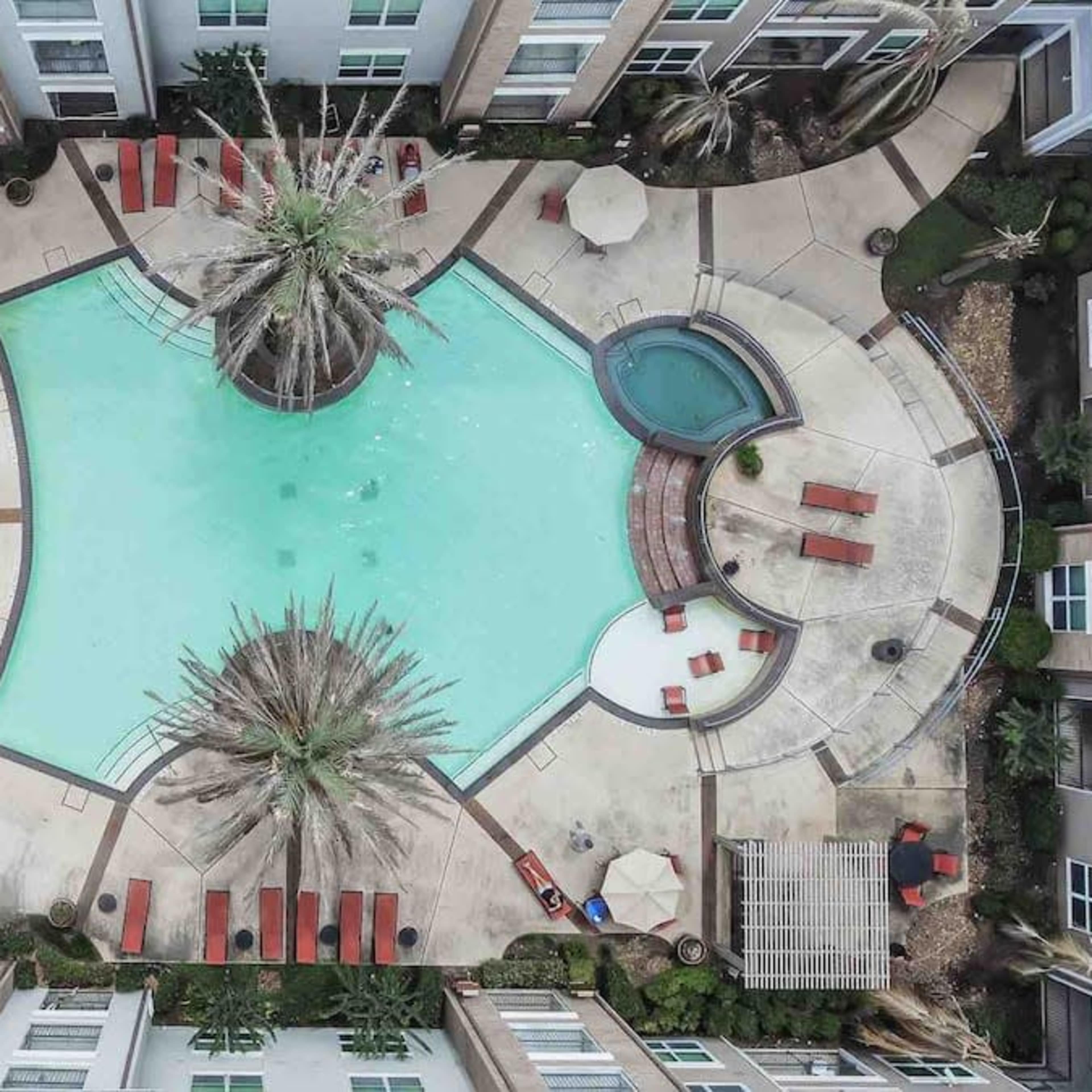 An aerial view of a swimming pool surrounded by lounge chairs and palm trees, located within a landscaped courtyard of an apartment complex.