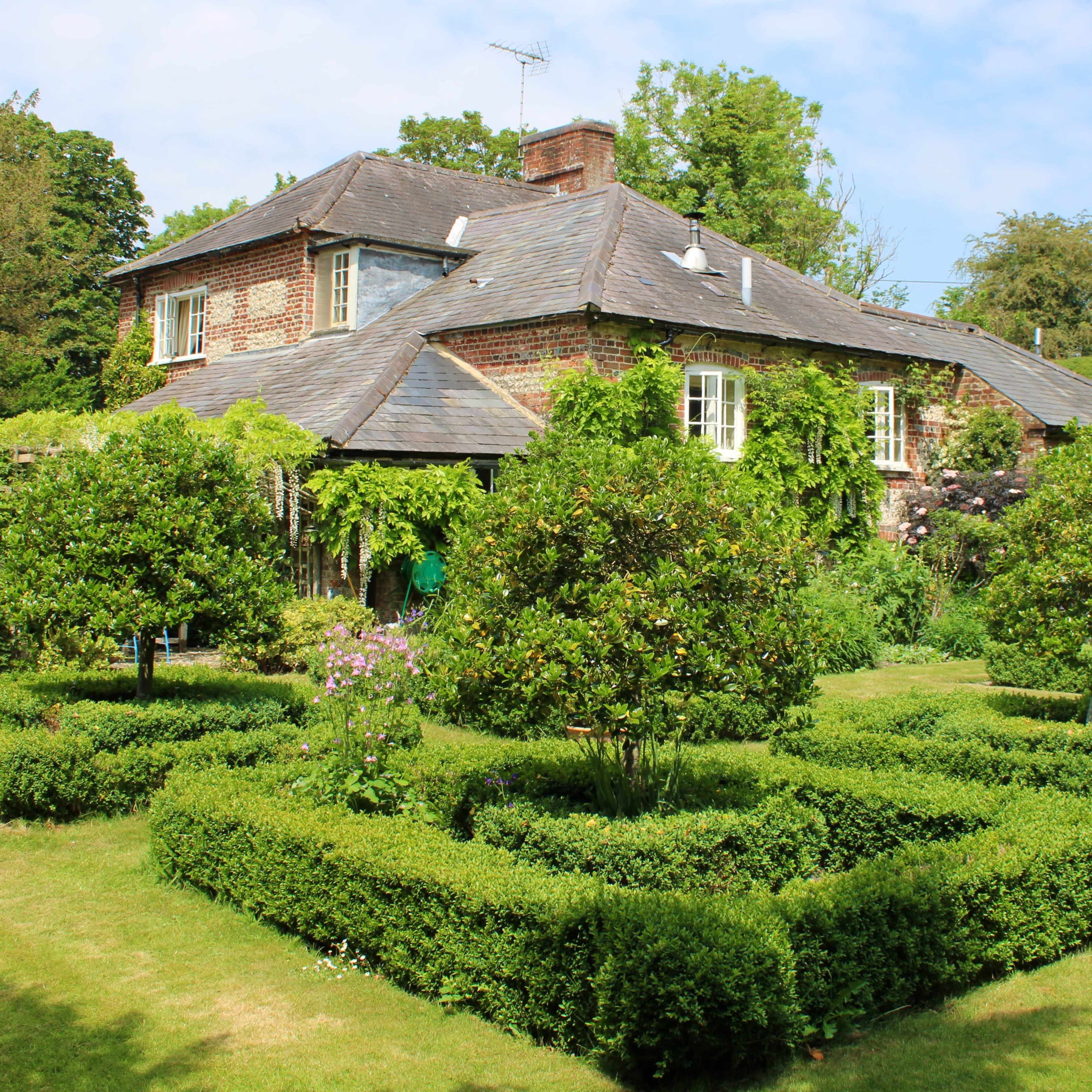 A brick house with a slate roof is surrounded by neatly trimmed hedges and small trees in a well-maintained garden.
