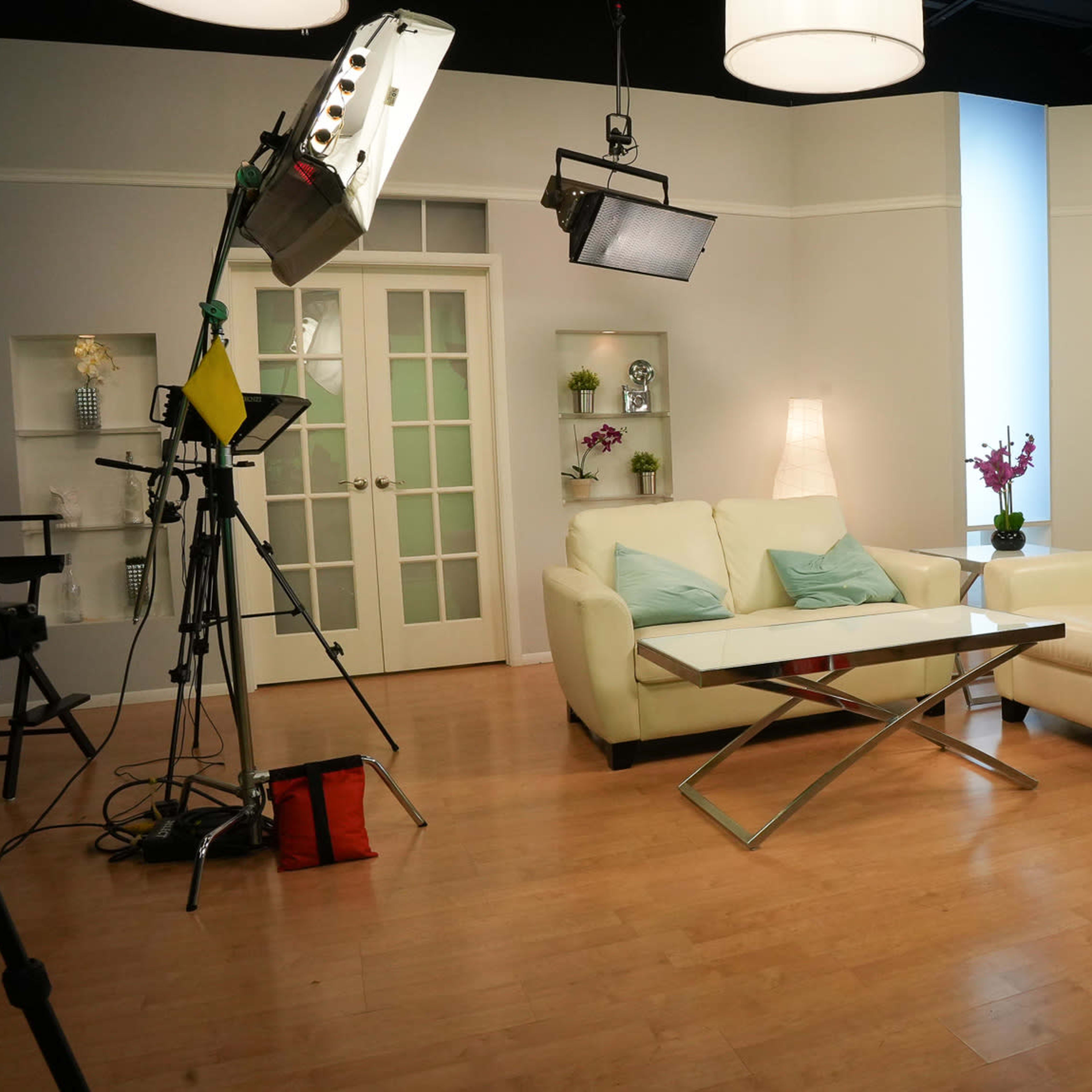 A well-lit television studio features a white sofa and armchair, a glass coffee table, and multiple camera setups.