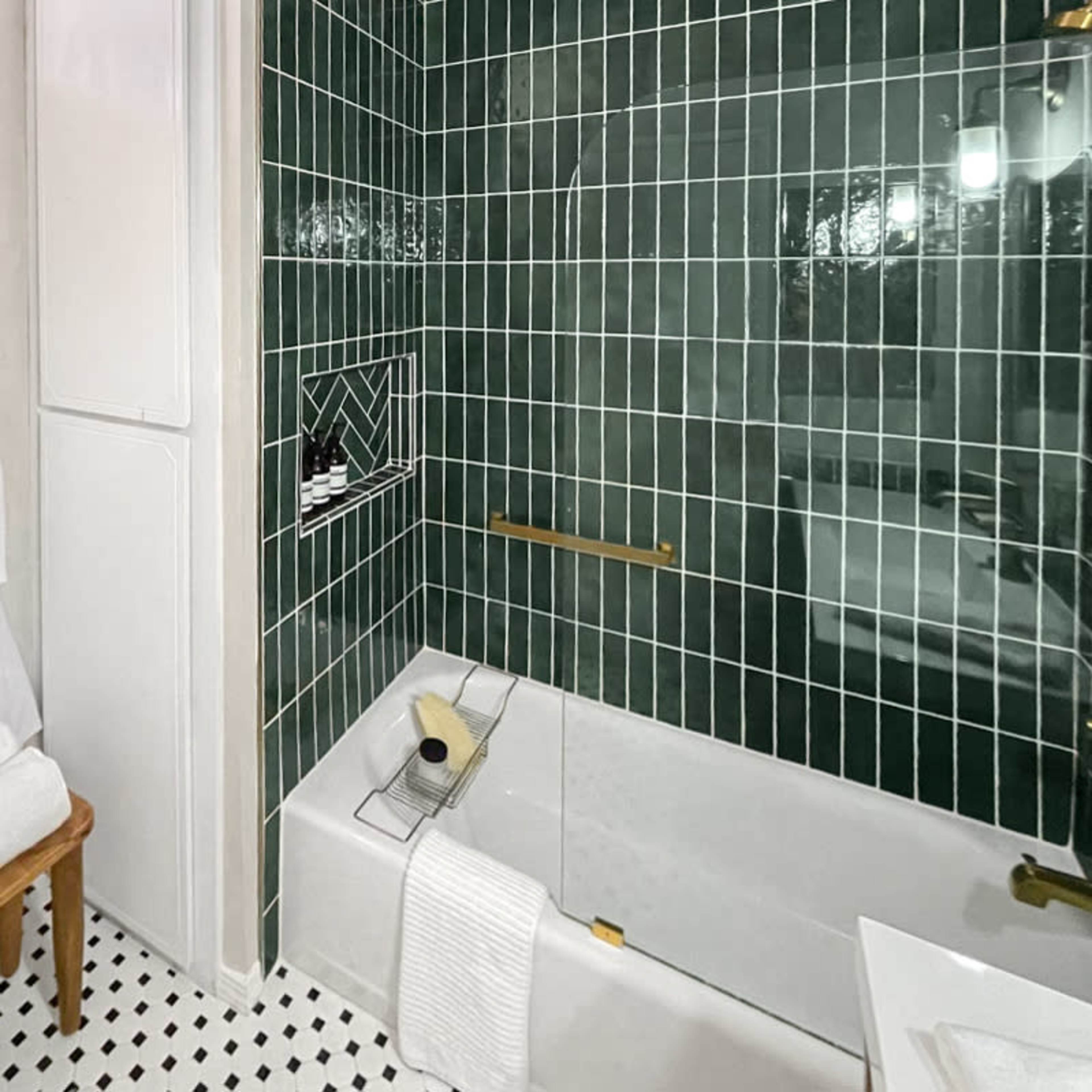 The bathroom features green tiled walls, a bathtub with a gold faucet, and neatly arranged white towels on a wooden stool.
