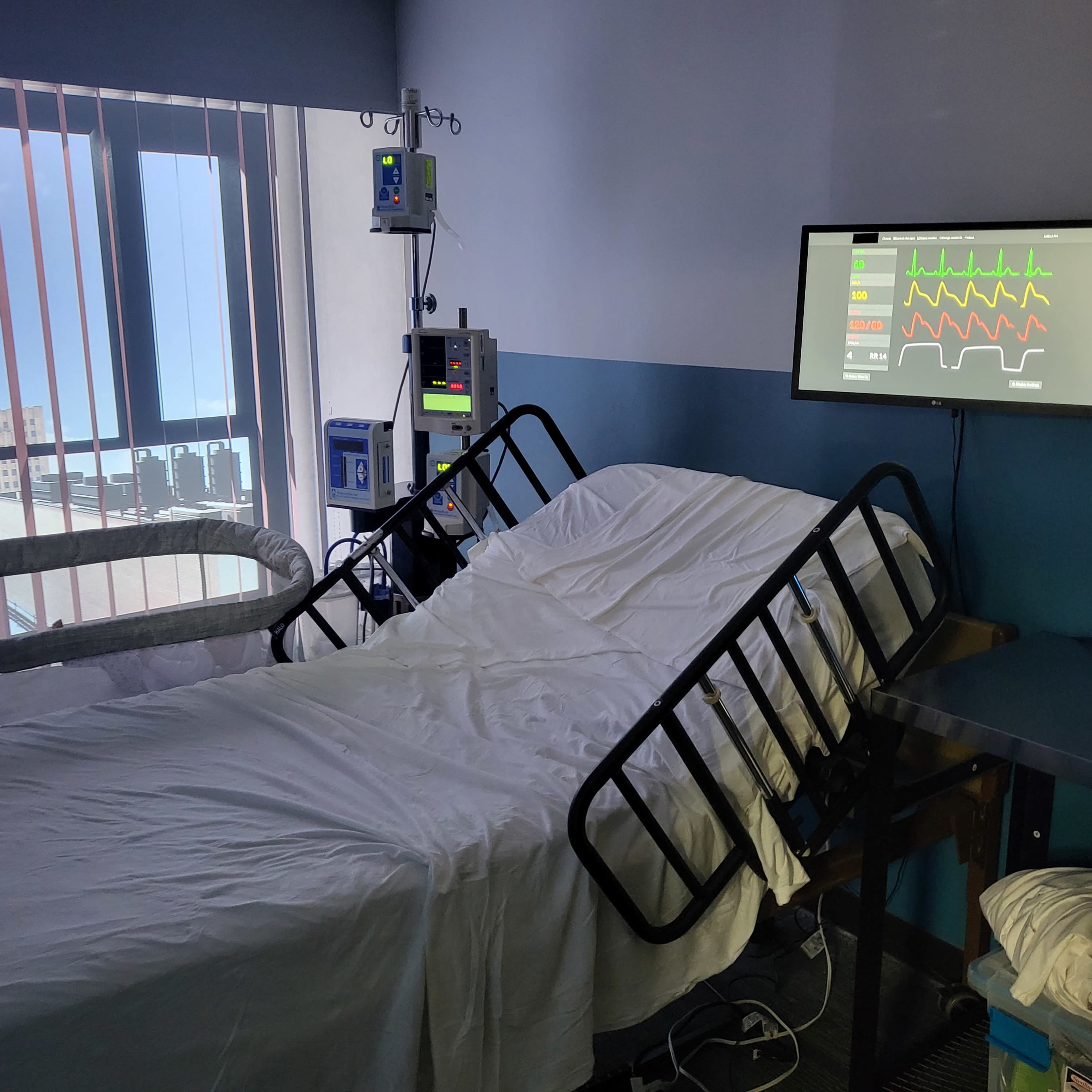 The room contains a hospital bed, medical monitoring equipment, and a laptop, with a large window letting in natural light.